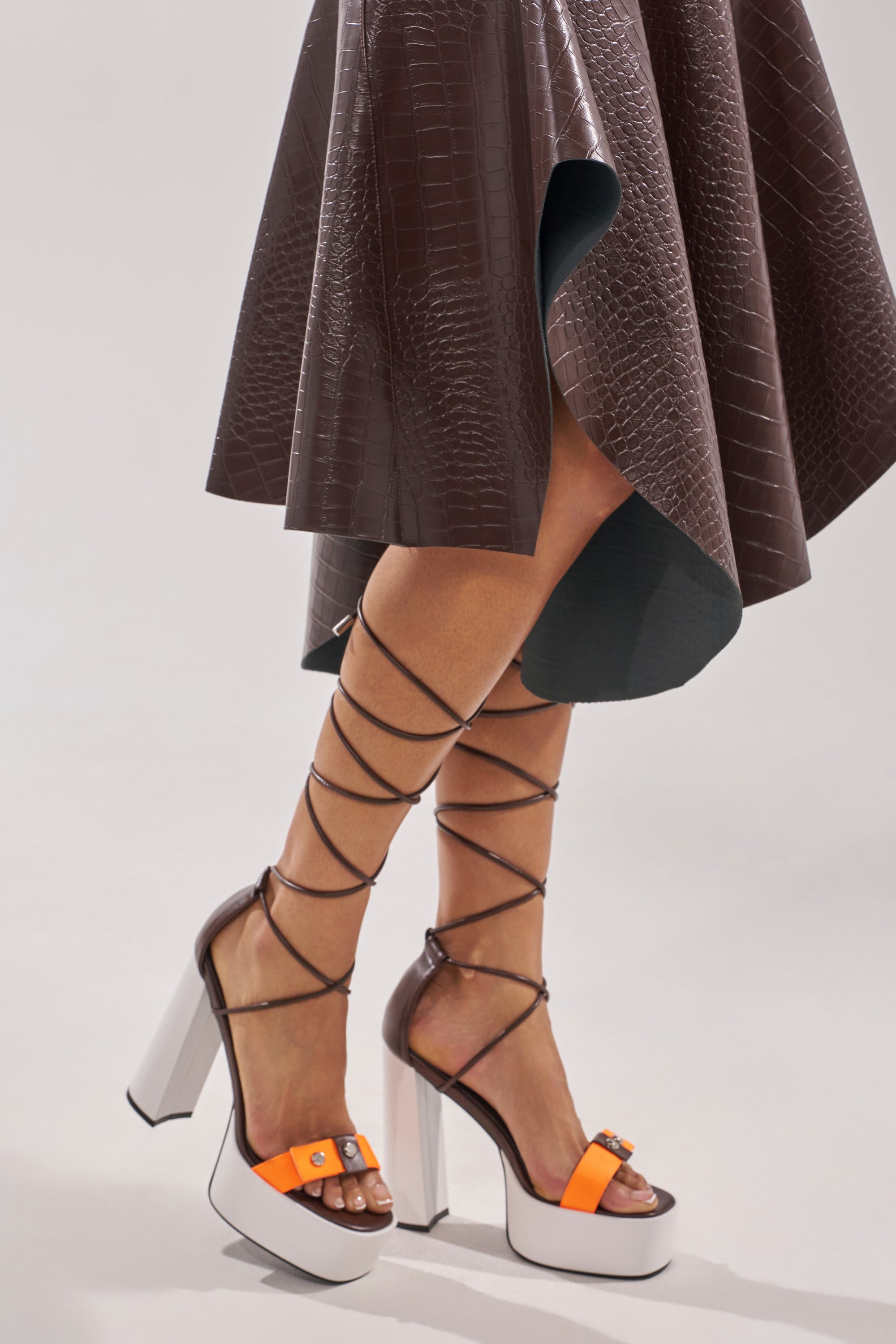 A person wears a brown, crocodile-patterned skirt and the AKIRA GOLD OREA ORANGE MULTI COLORBLOCKED PLATFORM SANDAL—high-heeled with orange straps, a block heel, and a chic tie-up design wrapping around the lower legs. 0