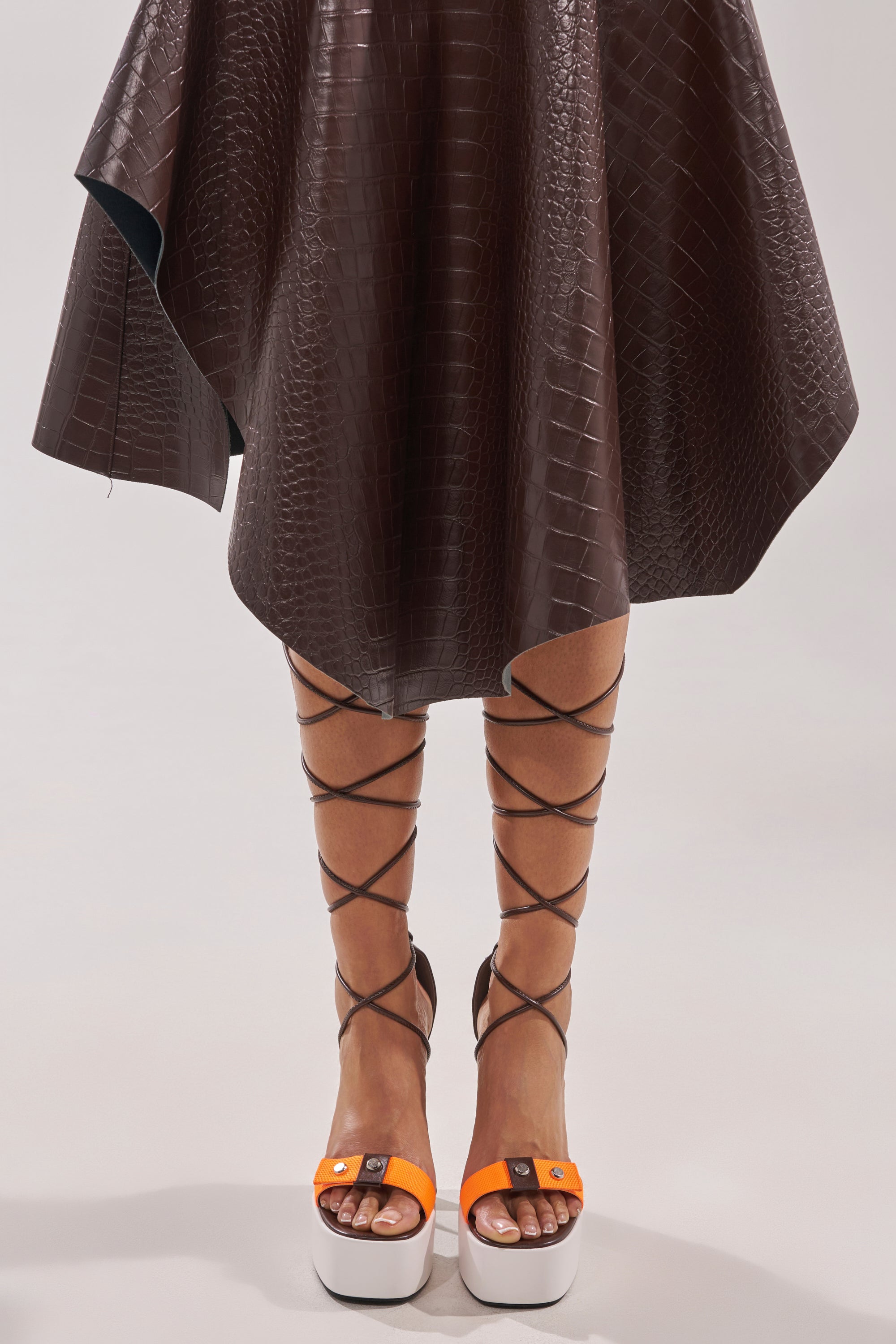 A person wearing a textured dark brown, knee-length skirt and AKIRA GOLD OREA ORANGE MULTI COLORBLOCKED PLATFORM SANDALS with block heels, tie-up black straps, white soles, and an eye-catching orange design. 1