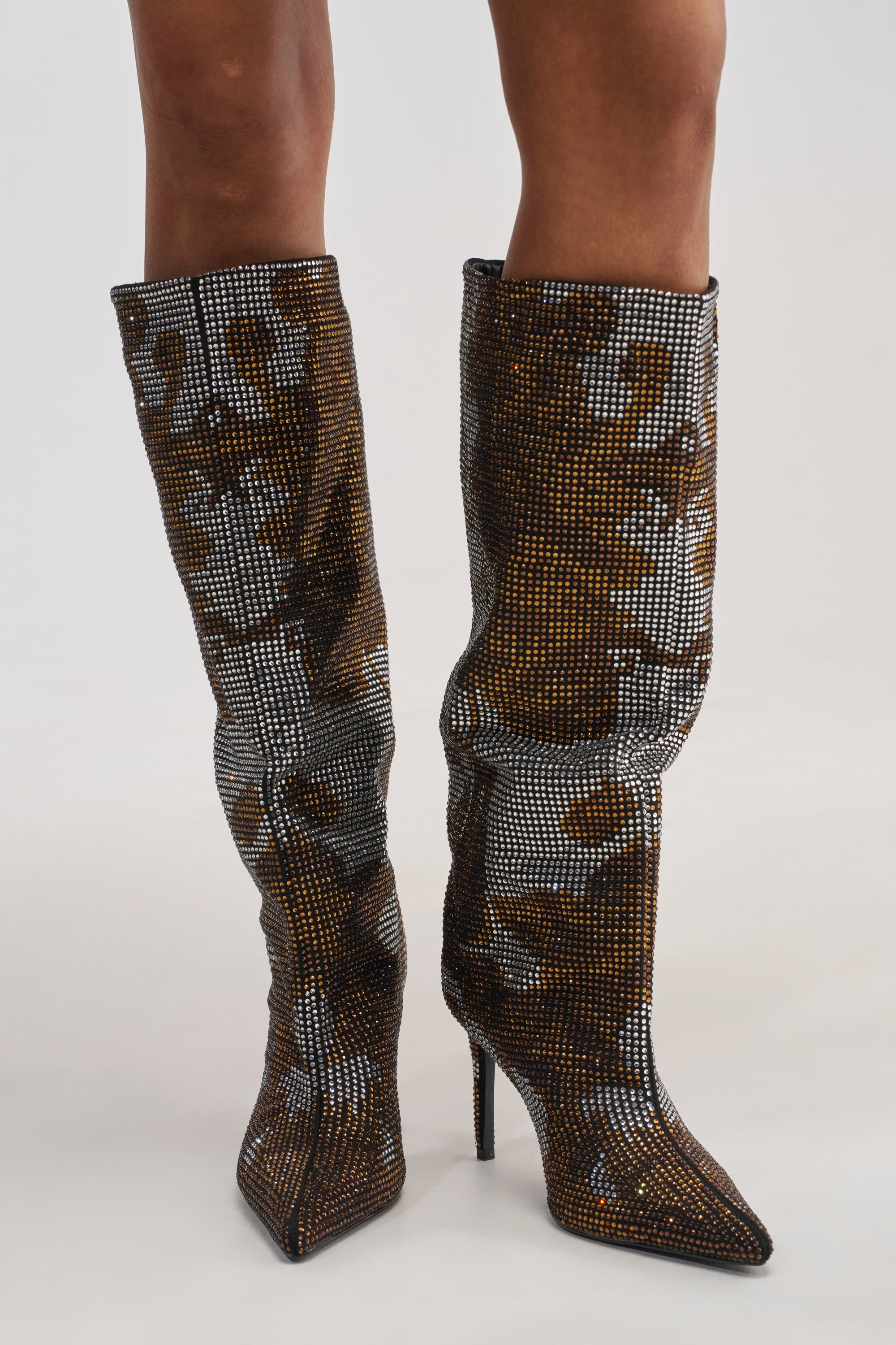 Wearing the AZALEA WANG ORIEL BRONZE MULTI PRINT RHINESTONE STILETTO BOOT—knee-high boots featuring a pointed toe, shimmering rhinestone embellishments, and a camouflage-inspired bronze, black, and silver pattern—exudes bold style worthy of attention.