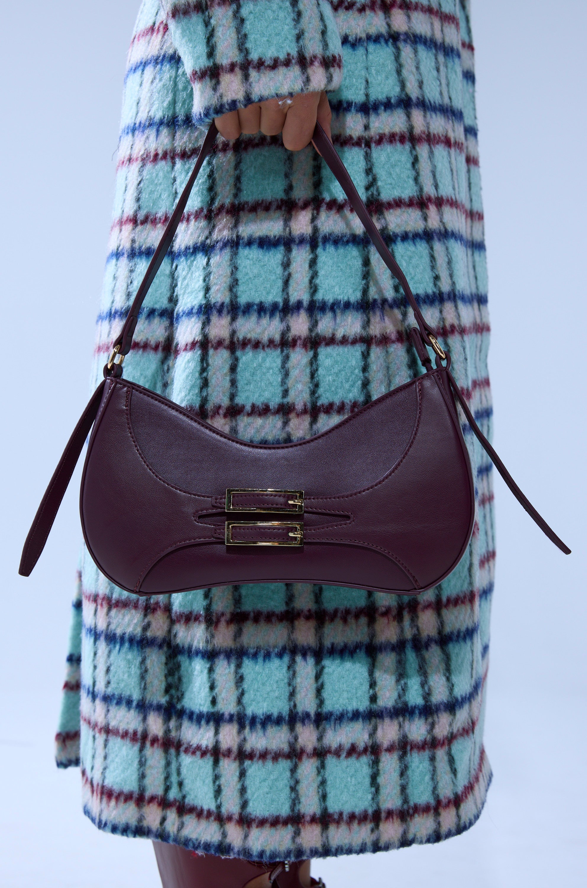 A person wearing a blue and red plaid coat holds the PLUM PURSE, a dark burgundy faux leather shoulder bag with gold-tone buckles and a shoulder strap. 1