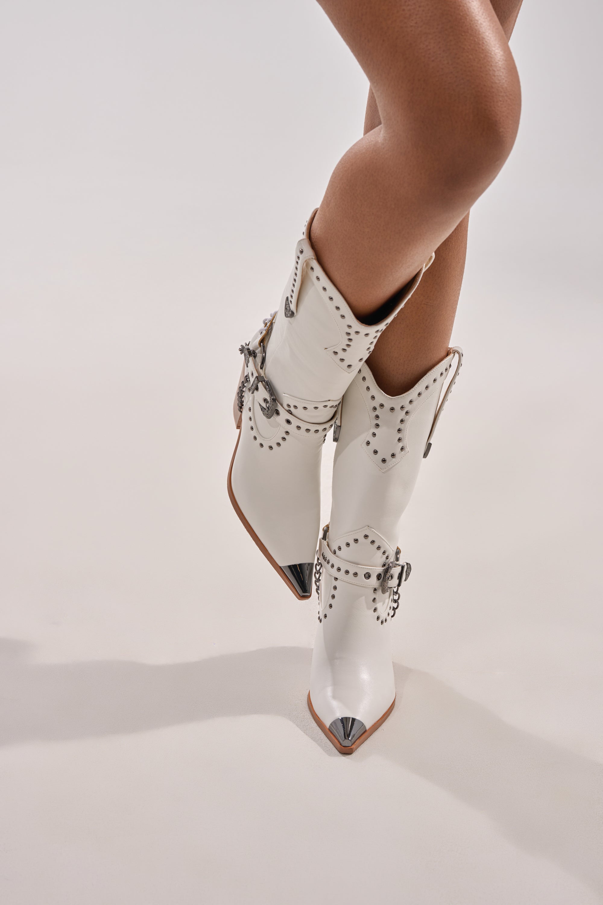 A person stands on a white surface, legs slightly crossed, wearing the AZALEA WANG OSBORN WHITE WESTERN BOOTIE featuring silver studs, decorative buckles, and pointed metallic toes.