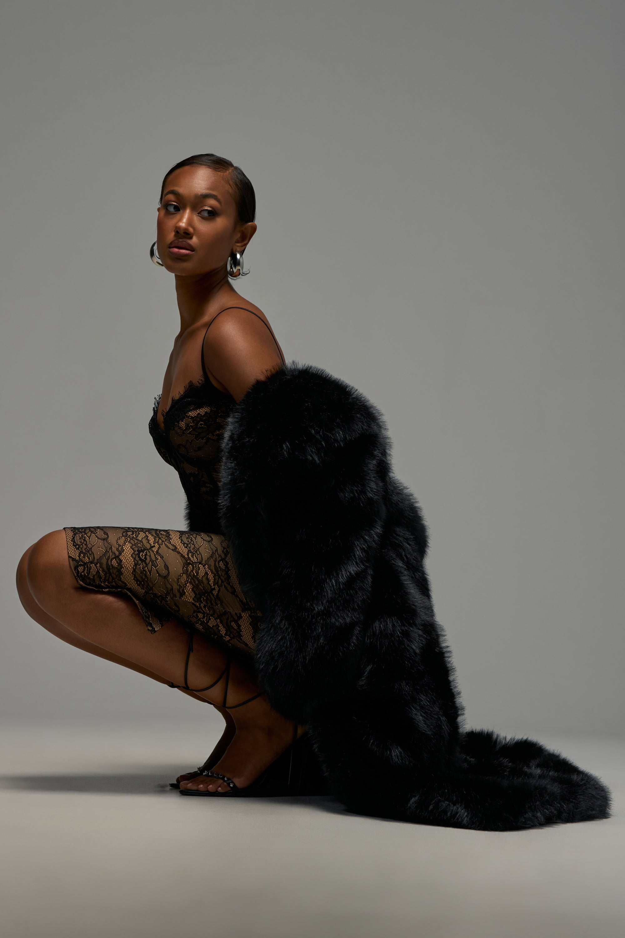 Draped in the OSLO FAUX FUR COAT IN BLACK over her shoulders, a woman kneels in a black lace dress and heels, accessorized with large hoop earrings for a winter-chic look against a plain gray background.