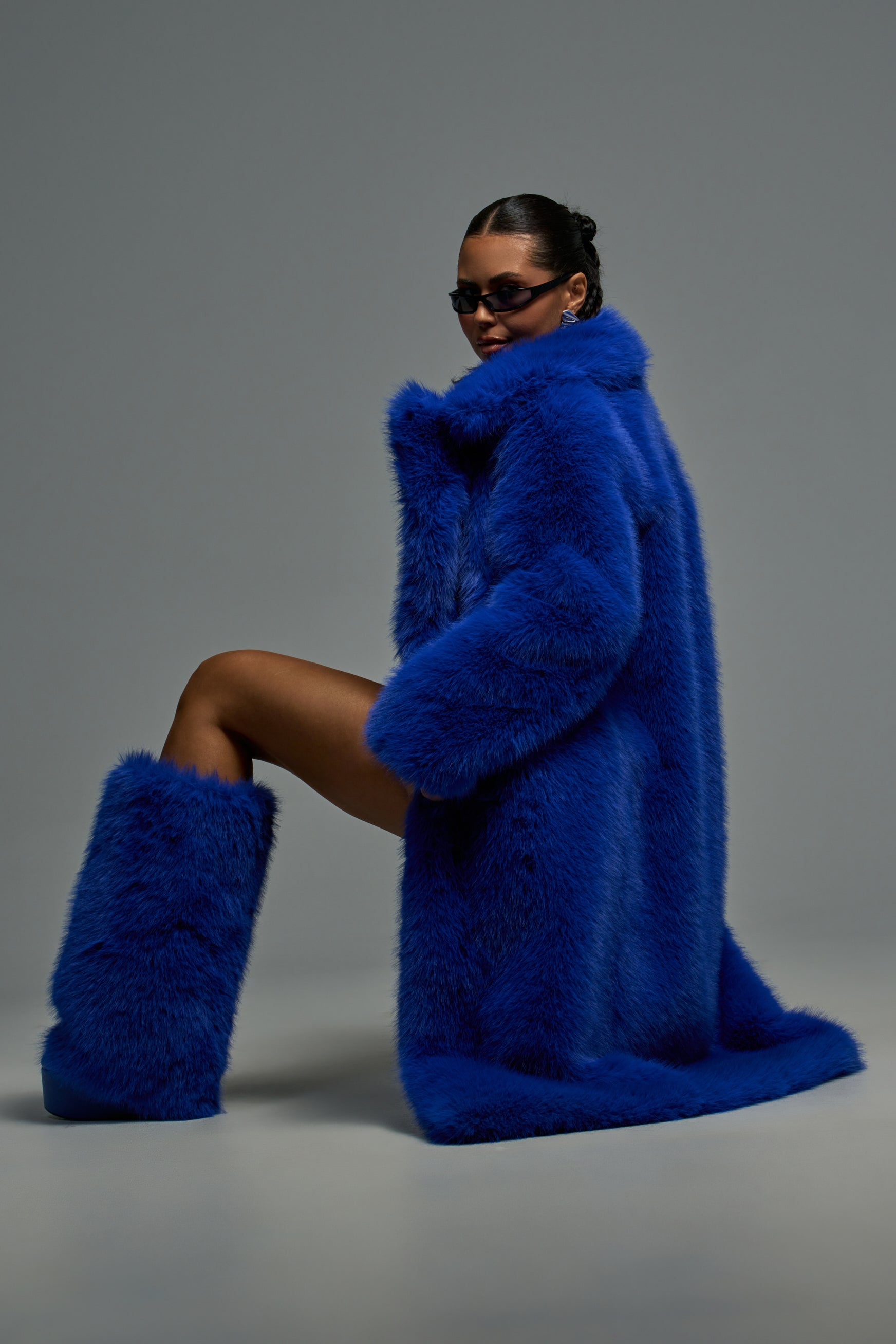 Wearing the OSLO FAUX FUR COAT IN COBALT and matching blue fur boots, a person poses against a plain gray background with their hair pulled back and dark sunglasses, showcasing bold cold weather style.