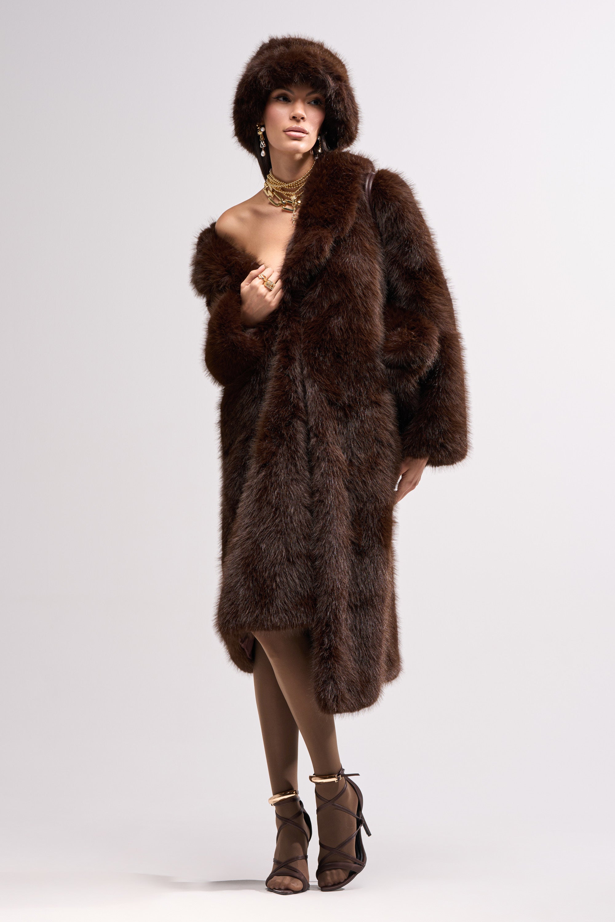 A woman wears the OSLO FAUX FUR COAT IN CHOCOLATE with a matching fur hat, sheer tights, and high heels. She accessorizes with gold necklaces and earrings, standing against a plain light background.