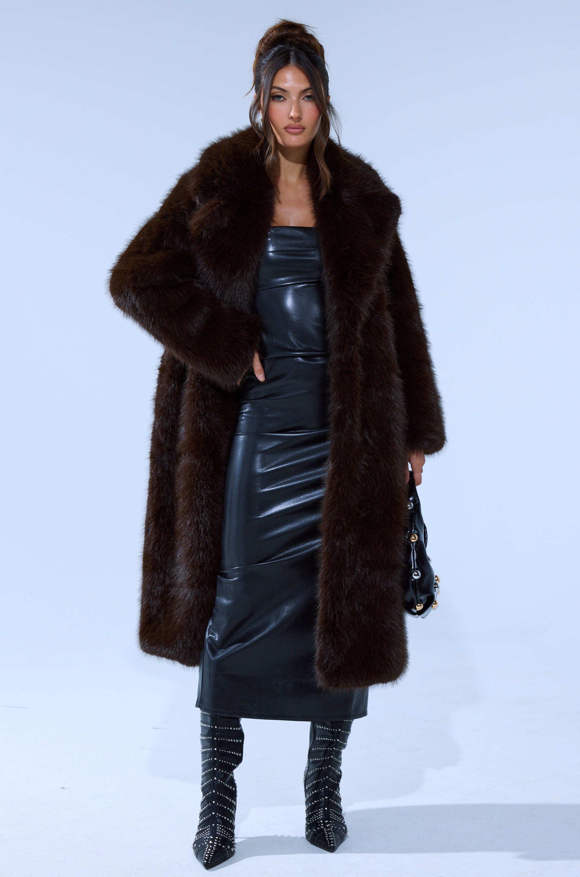 A woman poses in the OSLO FAUX FUR COAT IN CHOCOLATE over a shiny black leather dress, black studded knee-high boots, and holds a small black handbag against a plain light background.
