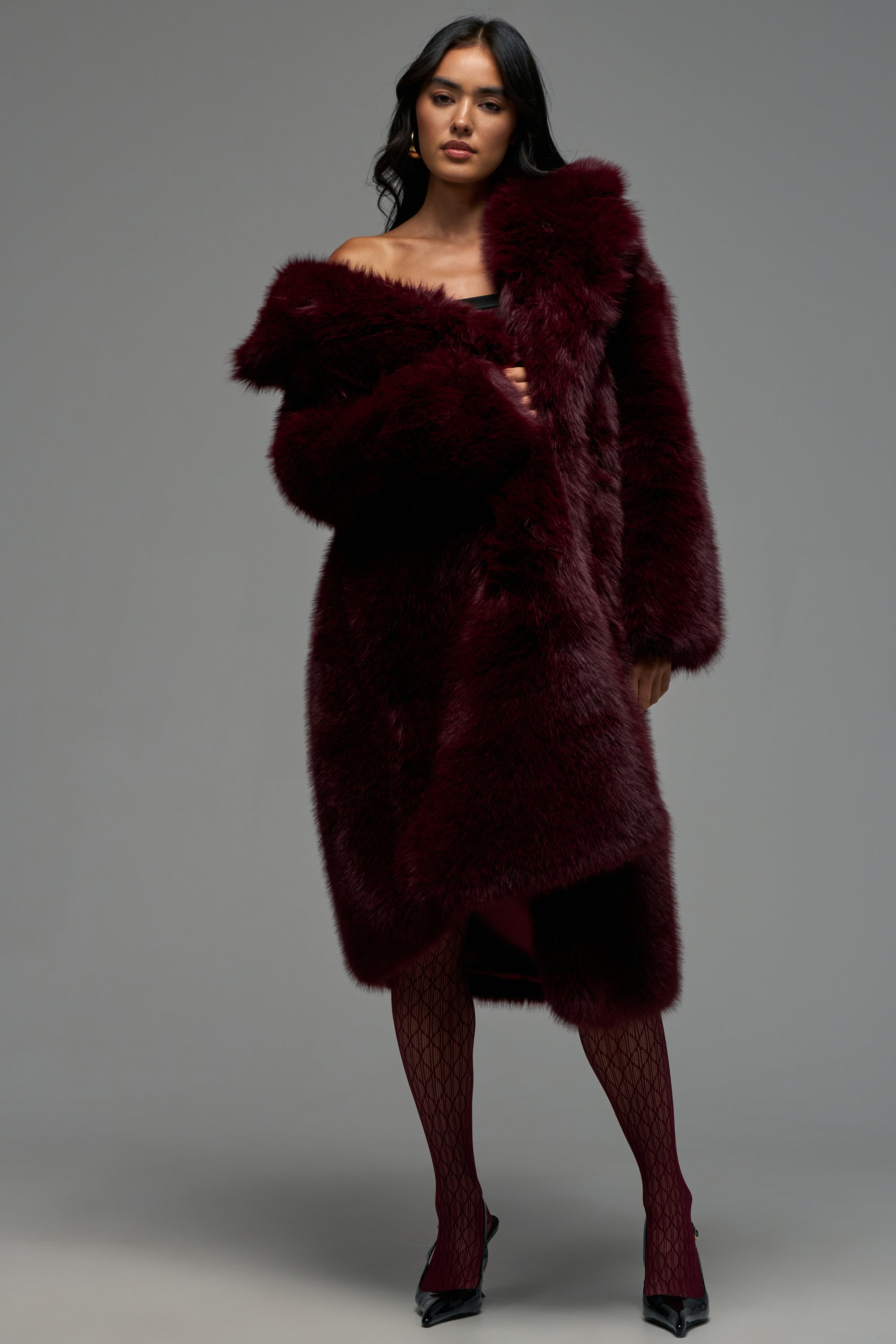 A woman poses in the OSLO FAUX FUR COAT IN BURGUNDY—a luxurious, deep burgundy longline faux fur coat—layered over a matching dress, styled with patterned tights and black heels, against a plain gray background.