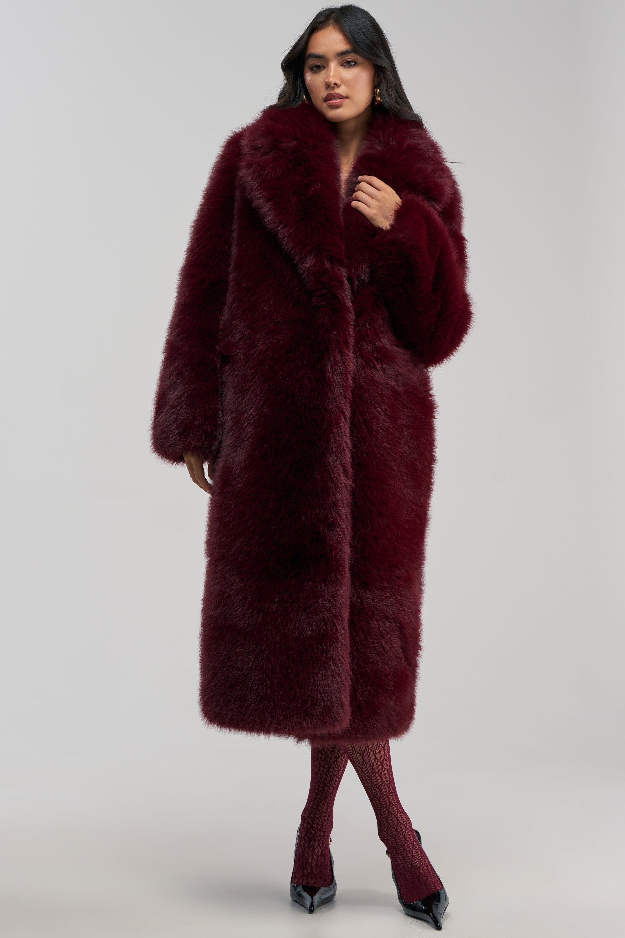 Dressed confidently in the OSLO FAUX FUR COAT IN BURGUNDY, paired with matching textured tights and black pointed shoes, a woman showcases impeccable winter styling against a plain background.
