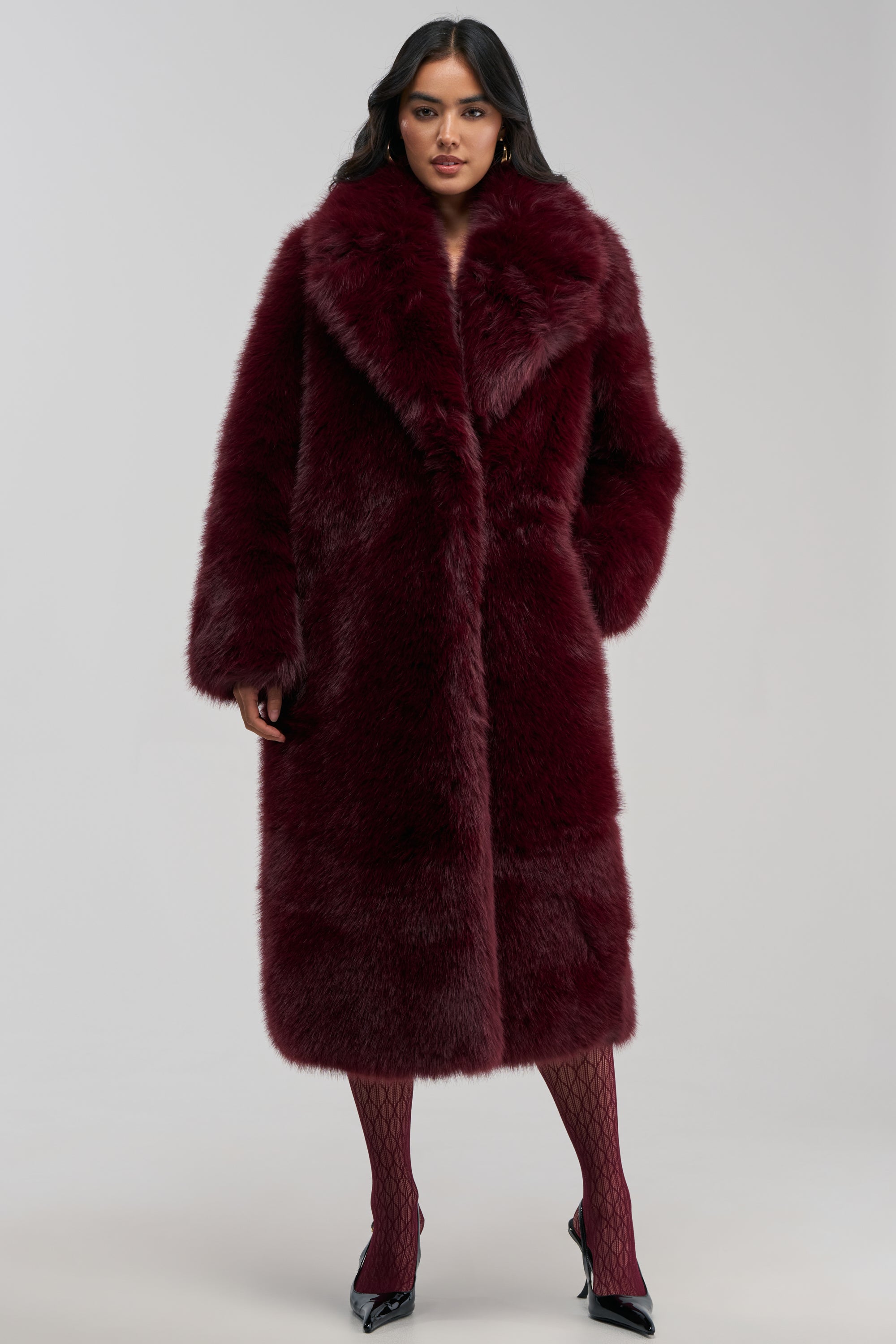 Wearing the OSLO FAUX FUR COAT IN BURGUNDY, this individual stands against a plain light background, exemplifying chic winter style with their long dark hair, patterned tights, and black pointed shoes.