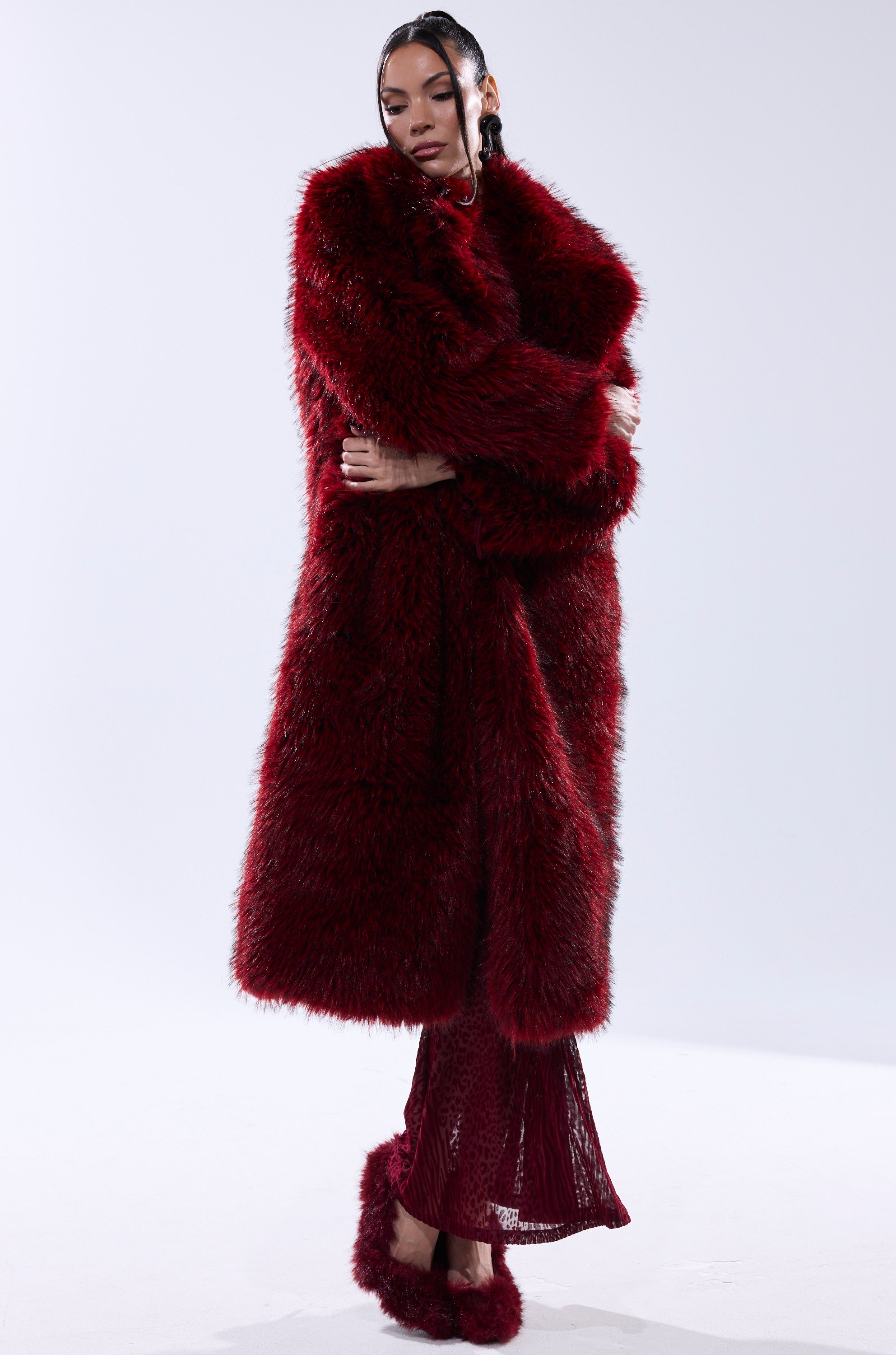 A person poses in the OSLO FAUX FUR COAT IN RED MULTI over a matching red dress and red fur slippers, arms crossed and head tilted against a plain white background.