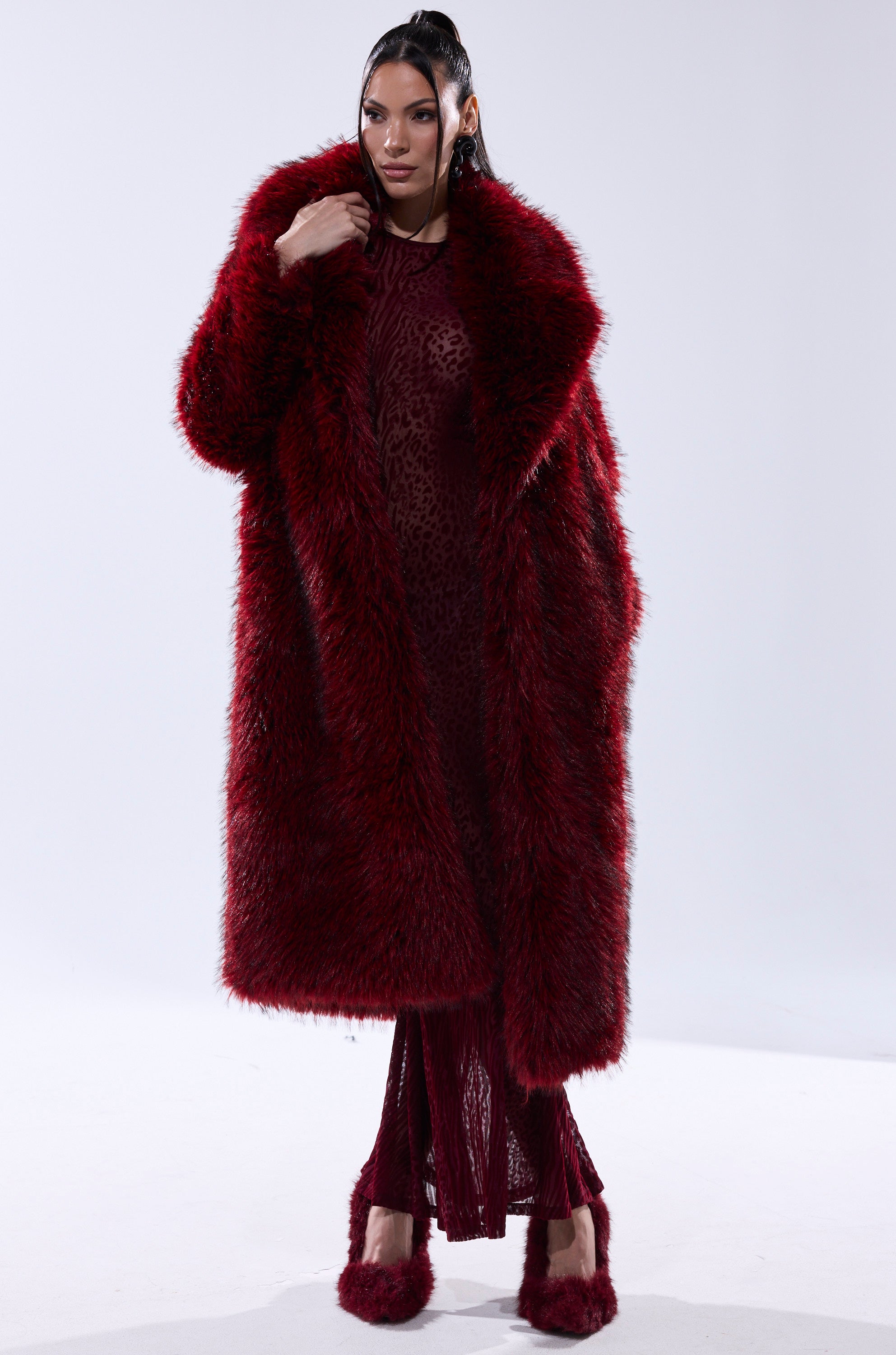 A woman stands in a studio wearing the OSLO FAUX FUR COAT IN RED MULTI over a matching patterned red outfit and red furry shoes. The background is plain and light-colored.