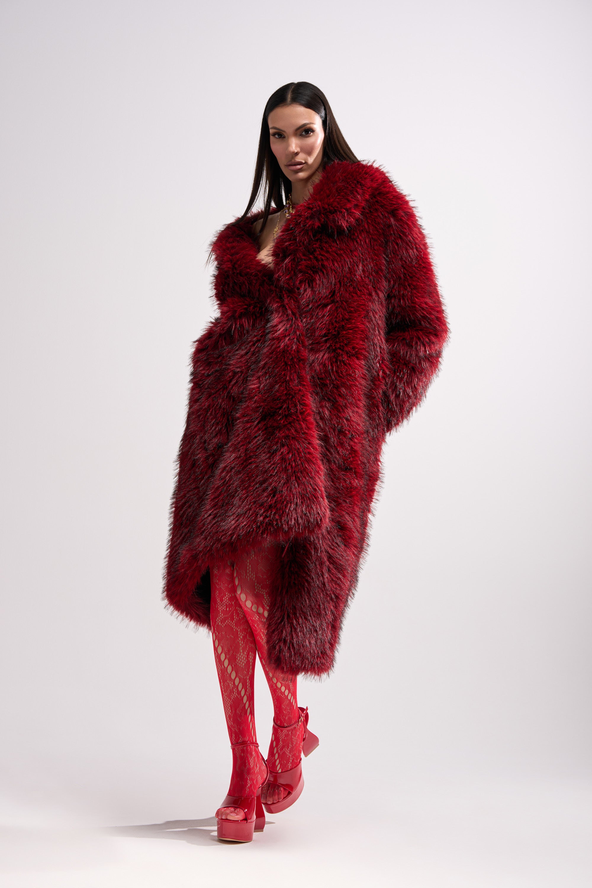 A woman exudes confidence in the OSLO FAUX FUR COAT IN RED MULTI, styled with red patterned tights and red platform heels against a simple light backdrop.