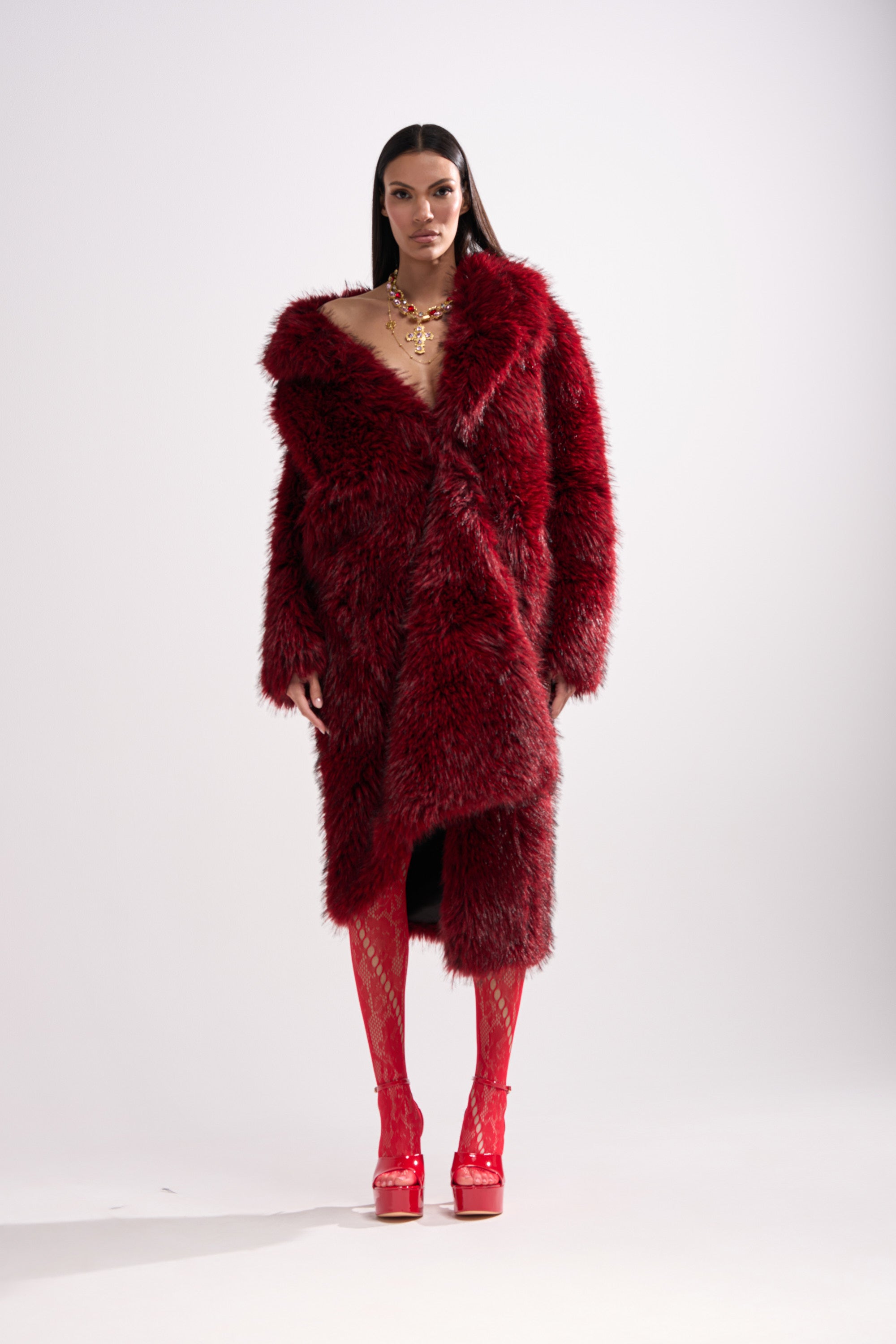 A woman with straight dark hair and a chunky gold necklace wears the OSLO FAUX FUR COAT IN RED MULTI against a plain backdrop, styled with red patterned tights and red platform heels.