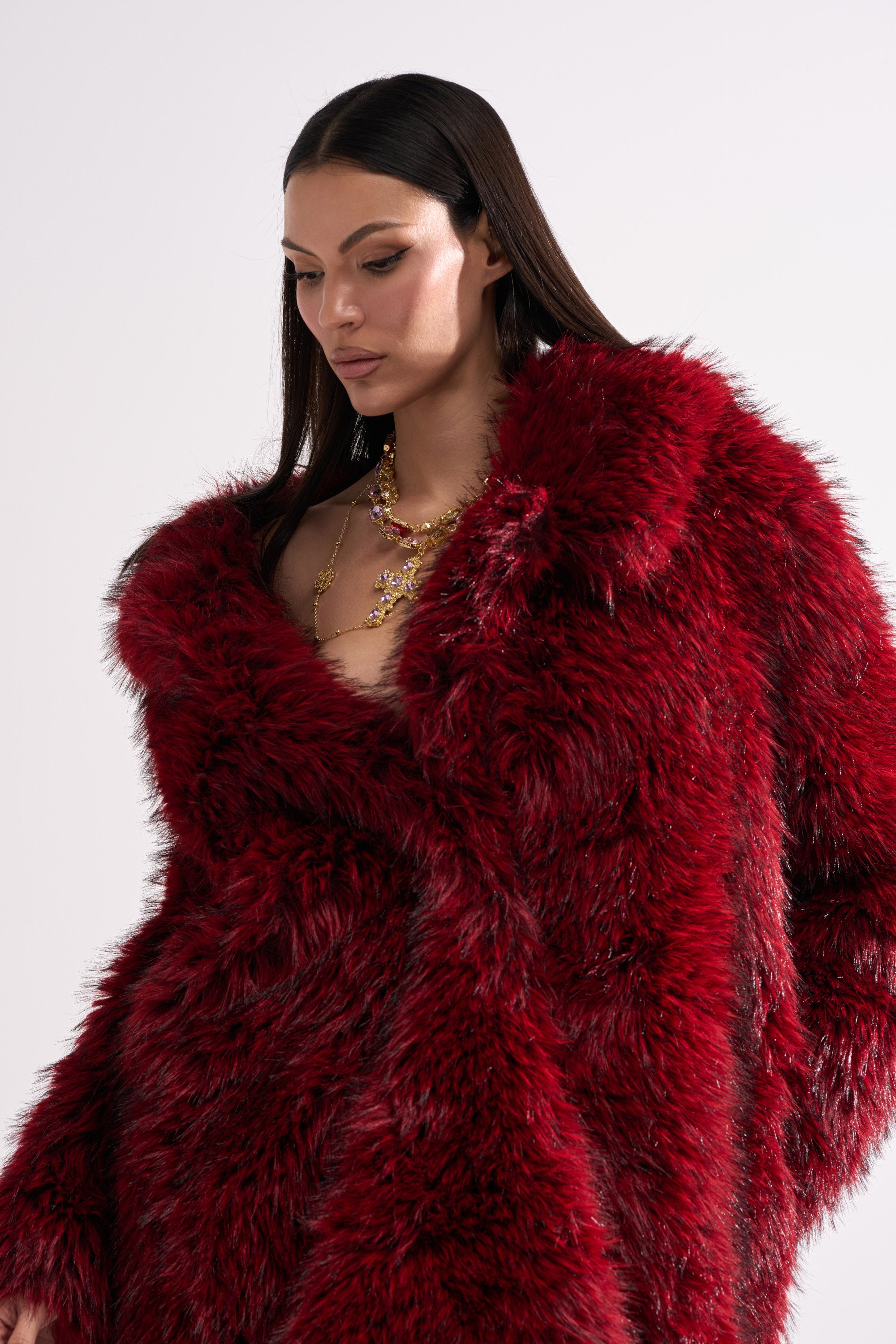 A woman with long dark hair wears the OSLO FAUX FUR COAT IN RED MULTI and a layered gold necklace while standing against a plain white background.