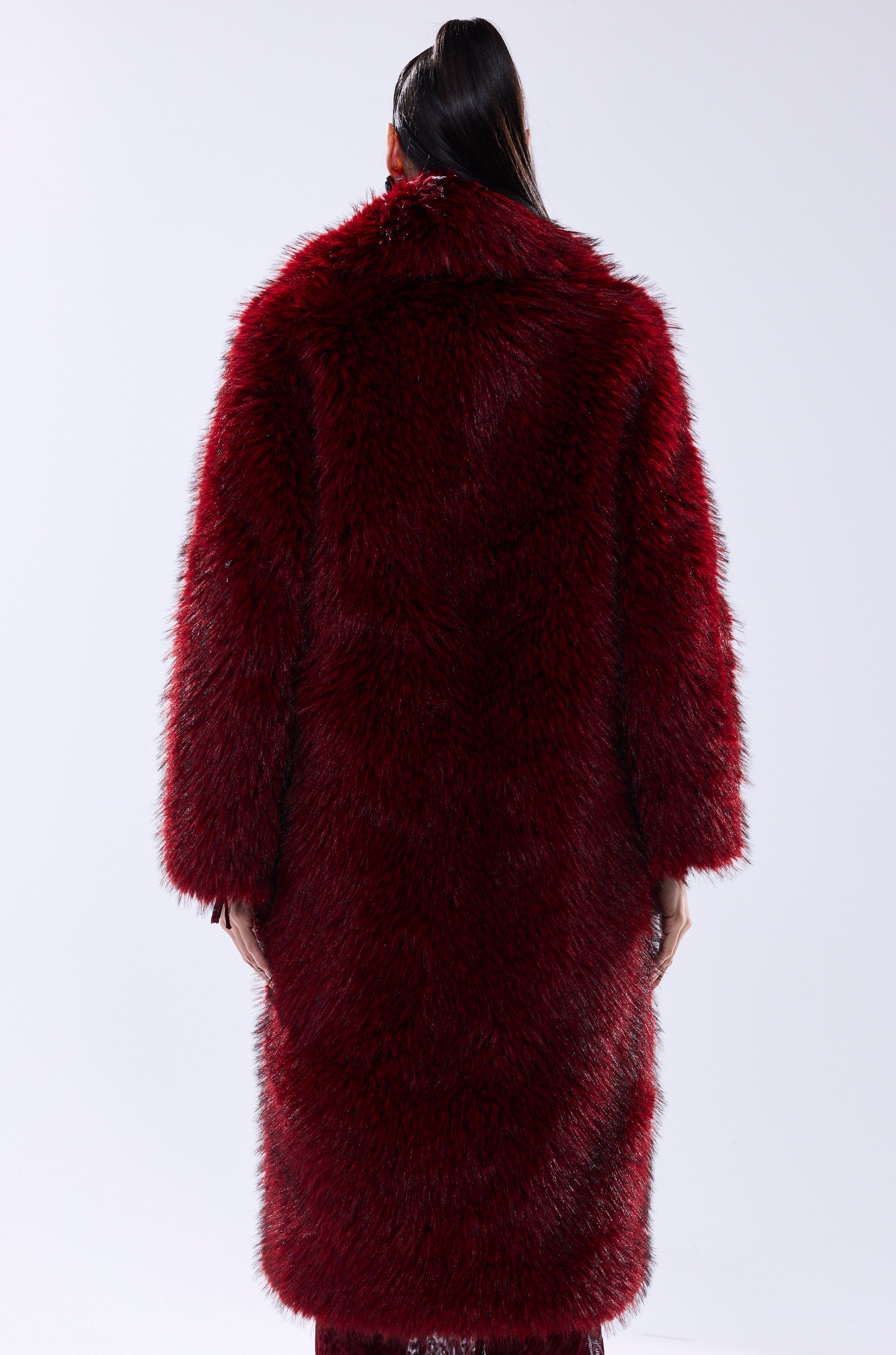 A person with long dark hair in a ponytail is seen from the back wearing the OSLO FAUX FUR COAT IN RED MULTI against a plain light gray background.