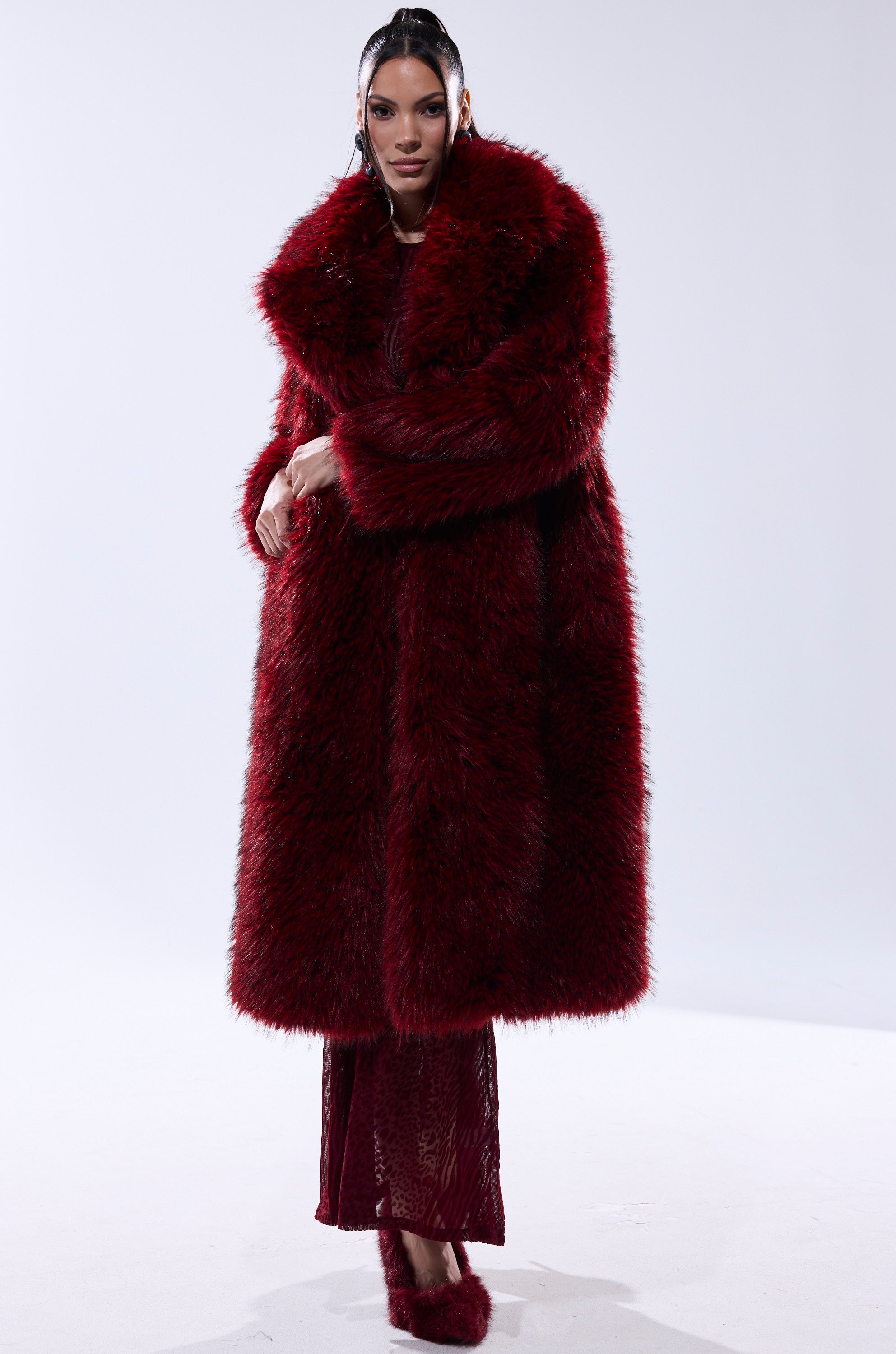 A woman with straight dark hair pulled back stands confidently against a plain background, wearing the OSLO FAUX FUR COAT IN RED MULTI over a matching red dress and red fur shoes.