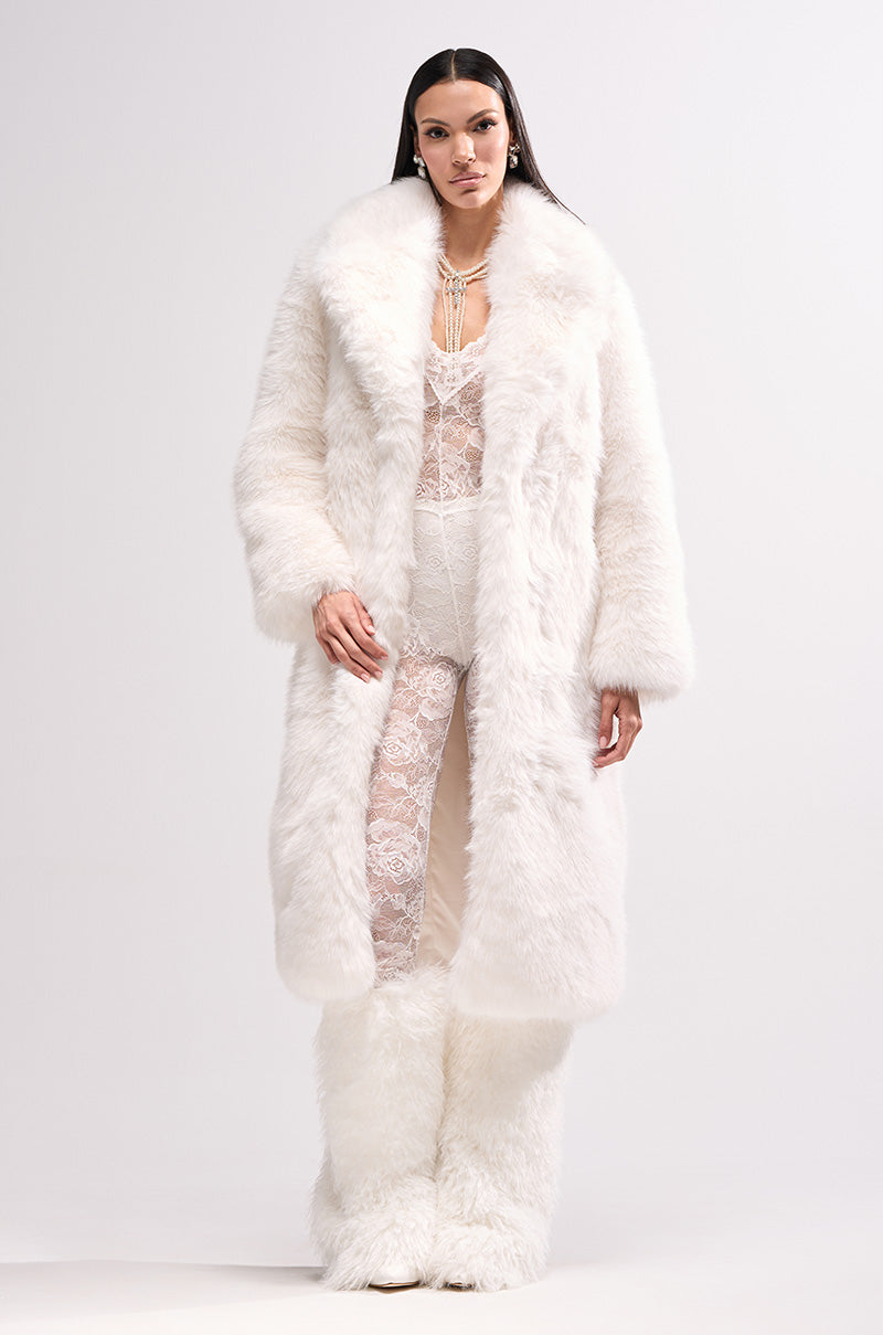 A woman poses in the OSLO FAUX FUR COAT IN WHITE, styled over a sheer white lace bodysuit and matching furry pants, standing against a light backdrop and looking directly at the camera.