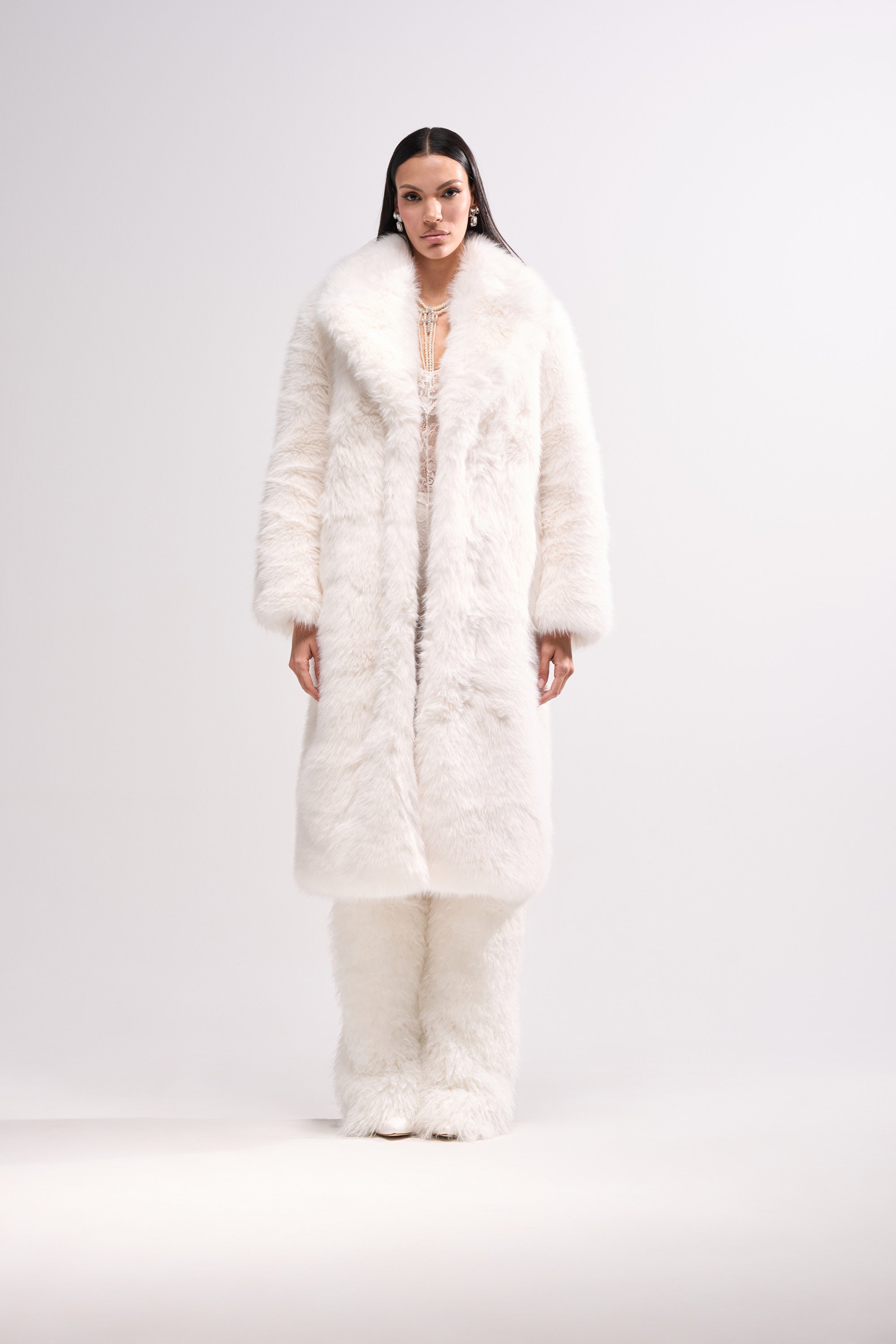 A person faces forward wearing the OSLO FAUX FUR COAT IN WHITE, styled with matching white fur pants against a plain light backdrop. The outfit appears warm and luxurious.