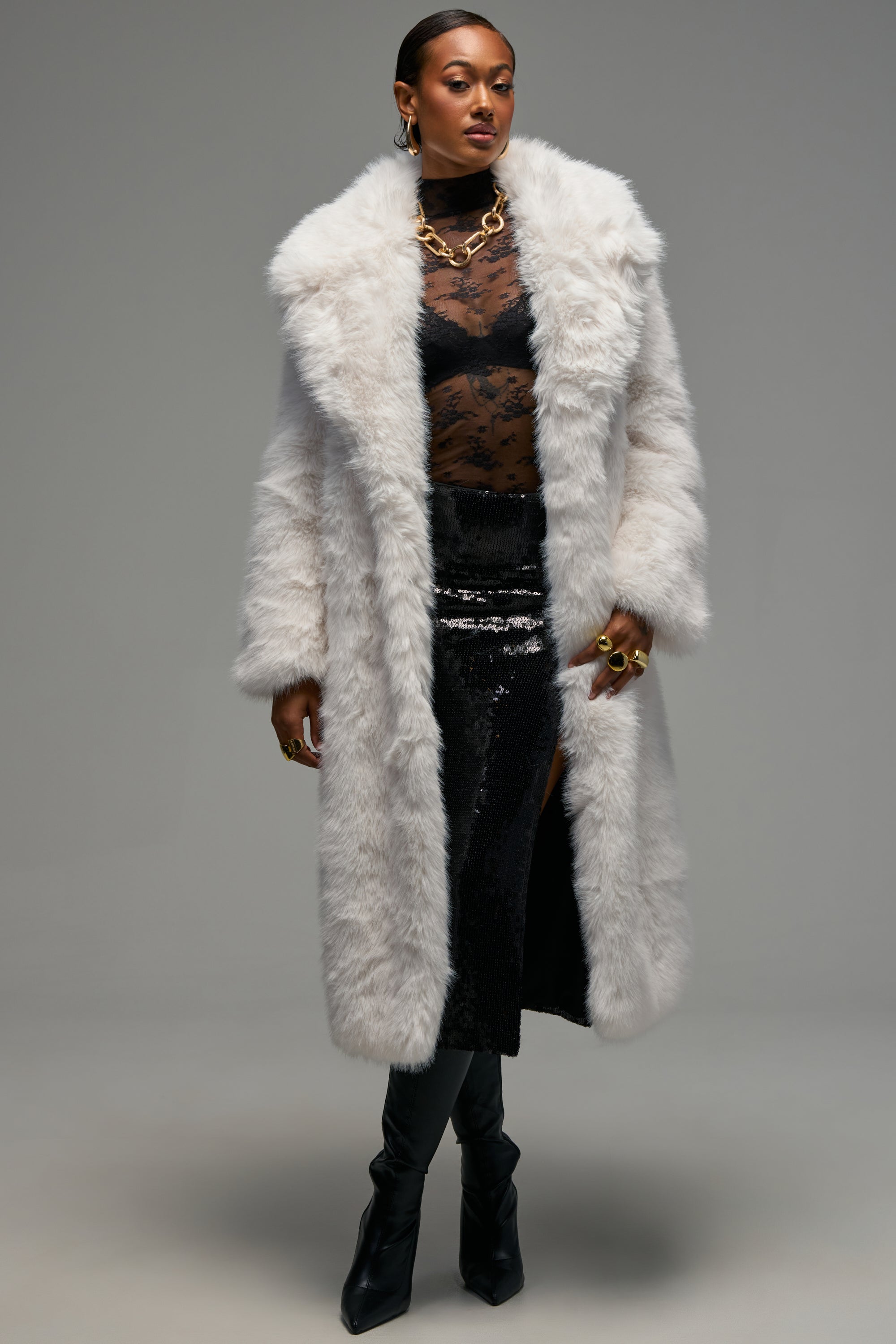 A woman stands against a plain gray background, exuding winter-chic style in the OSLO FAUX FUR COAT IN WHITE. She layers the luxurious coat over a sheer black lace top and black sequin skirt with a slit, completing her look with black boots, chunky gold jewelry, and gold rings.