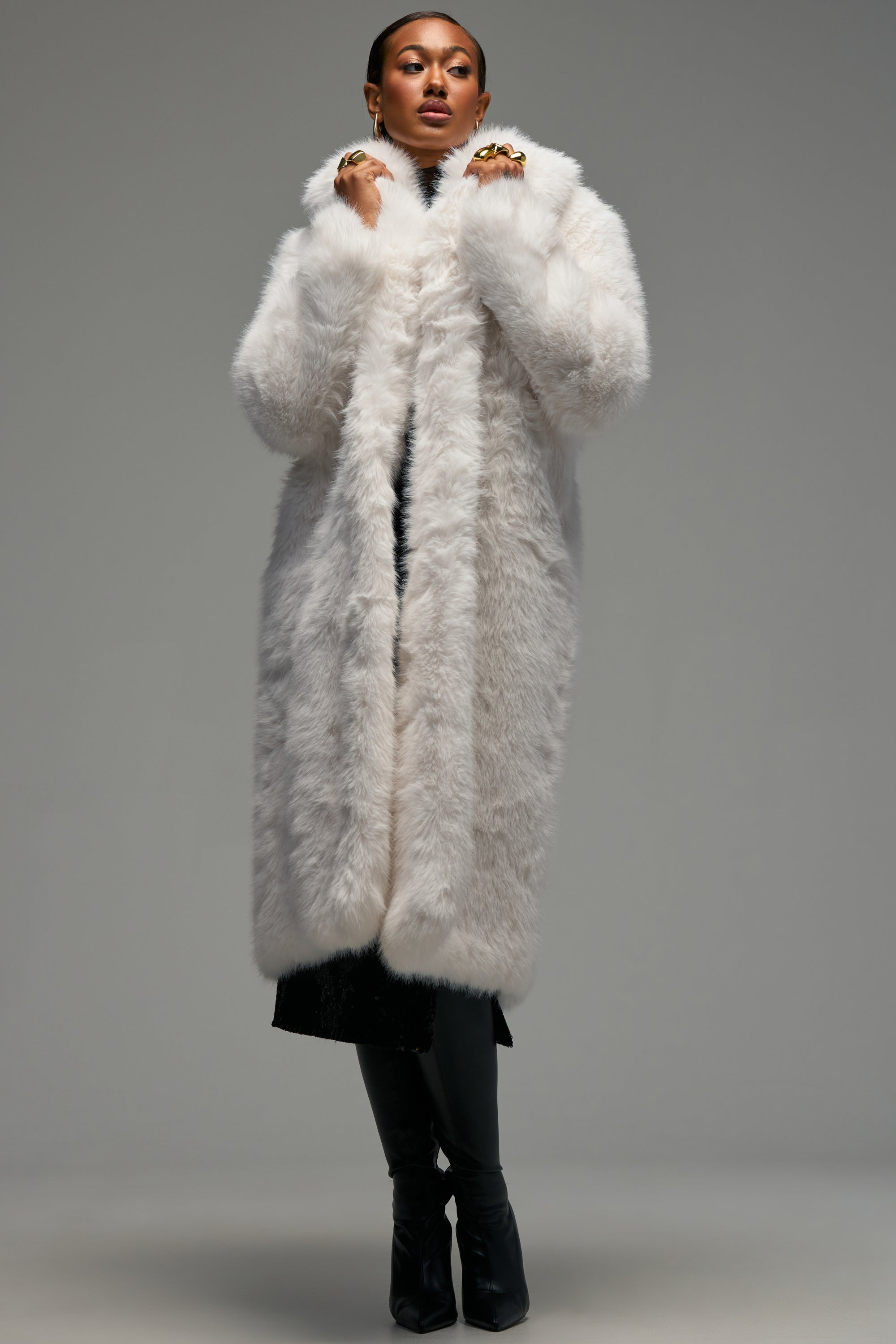 Dressed in the OSLO FAUX FUR COAT IN WHITE, a person stands confidently with opulent layers draped over black pants and boots. Hands lifted near their face highlight an array of rings, set against a simple gray background and radiating winter-chic allure.