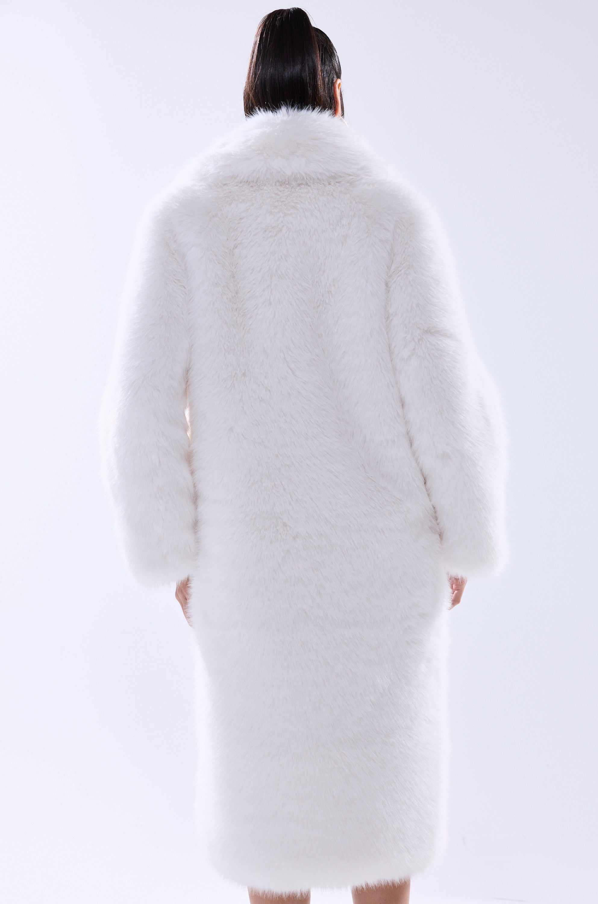 A person with long dark hair tied back stands with their back to the camera, wearing the OSLO FAUX FUR COAT IN WHITE—an oversized coat that falls below the knees. The background is plain and light.