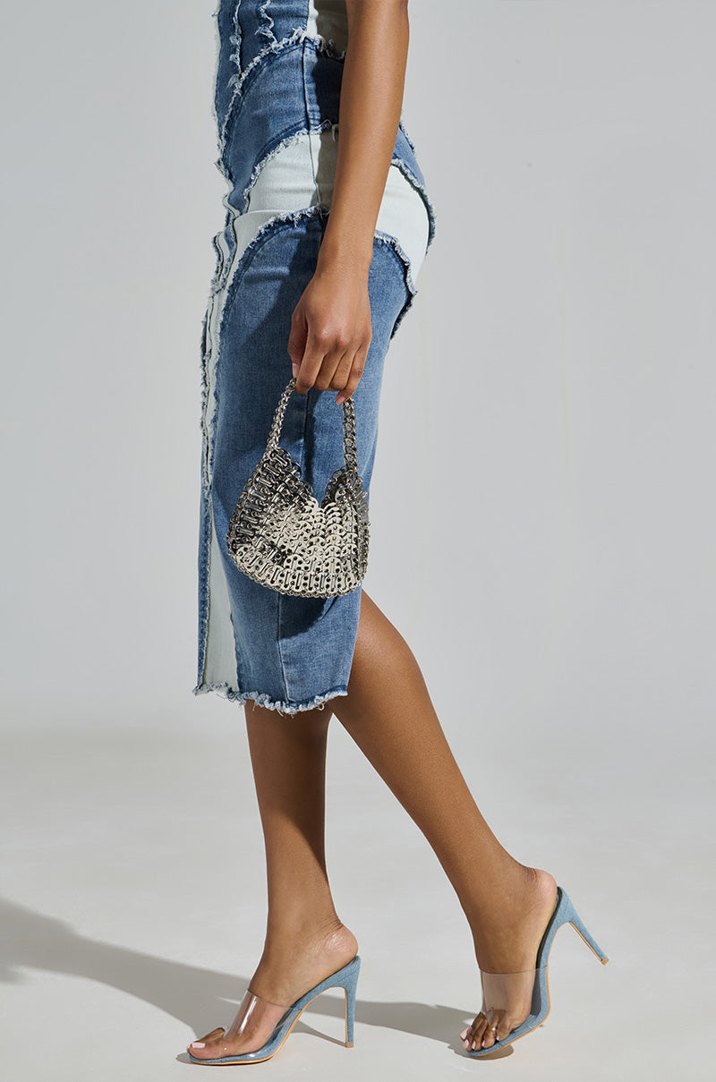 A person is wearing a two-tone denim skirt paired with AKIRA OSWEGO DENIM NEW SLIP ON MULE SANDALS, known for their comfort cushion insole. They hold a small woven metallic handbag against a plain light gray background, highlighting the chic outfit and stylish accessories.