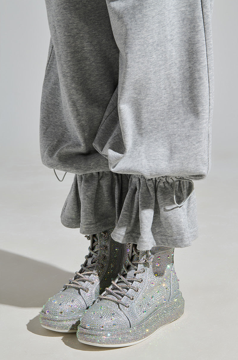 A person in gray sweatpants with ruffled cuffs completes their look with AZALEA WANG Otis Rhinestone High Top Sneakers in silver, standing on a light gray floor.