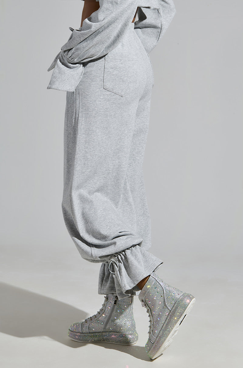 A person in loose-fitting gray sweatpants with tied ankle cuffs stands on a light gray background, wearing AZALEA WANG OTIS Rhinestone High Top Sneakers in Silver with thick soles, glittering under the light as they cross their arms casually.