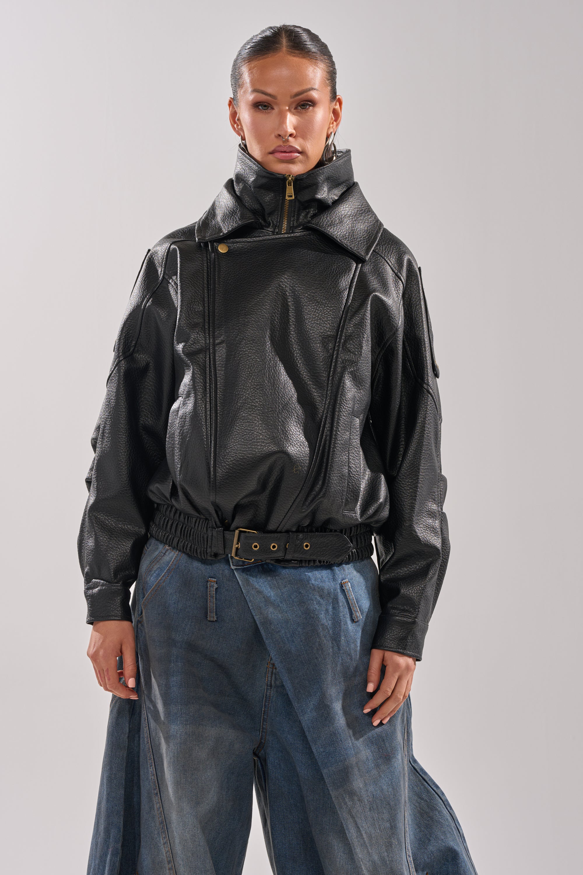 A woman with slicked-back hair and a confident expression faces forward, wearing the ERIKKA OVERSIZED MOTO jacket with a biker collar and snap buttons, paired with loose wide-leg blue jeans and a black belt.