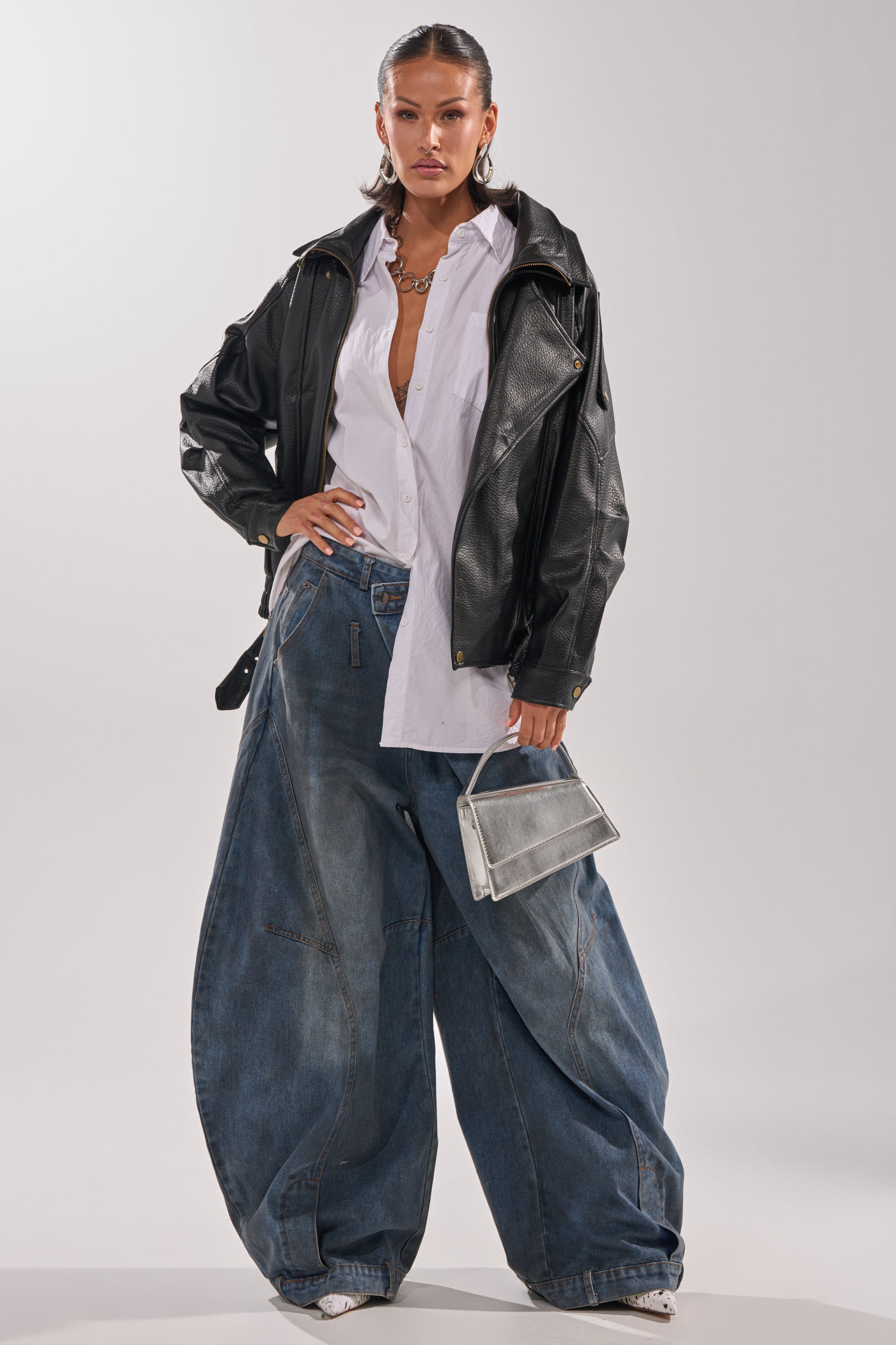 Wearing the ERIKKA OVERSIZED MOTO with a biker collar over a white shirt, she pairs it with baggy blue jeans and white shoes. A small silver handbag, hoop earrings, and a necklace complete her look against a plain backdrop.