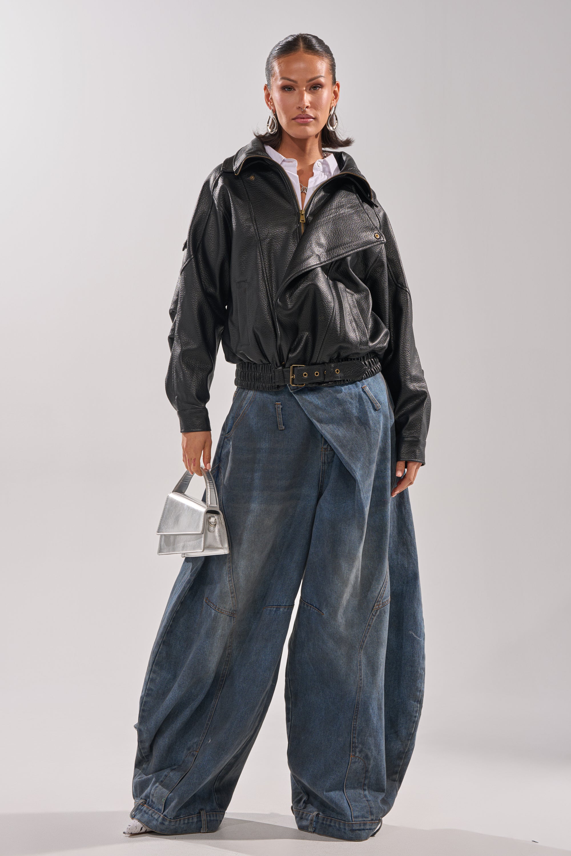 A woman poses against a plain light background wearing the ERIKKA OVERSIZED MOTO jacket, loose wide-leg blue jeans, white heeled shoes, and holding a small silver handbag.