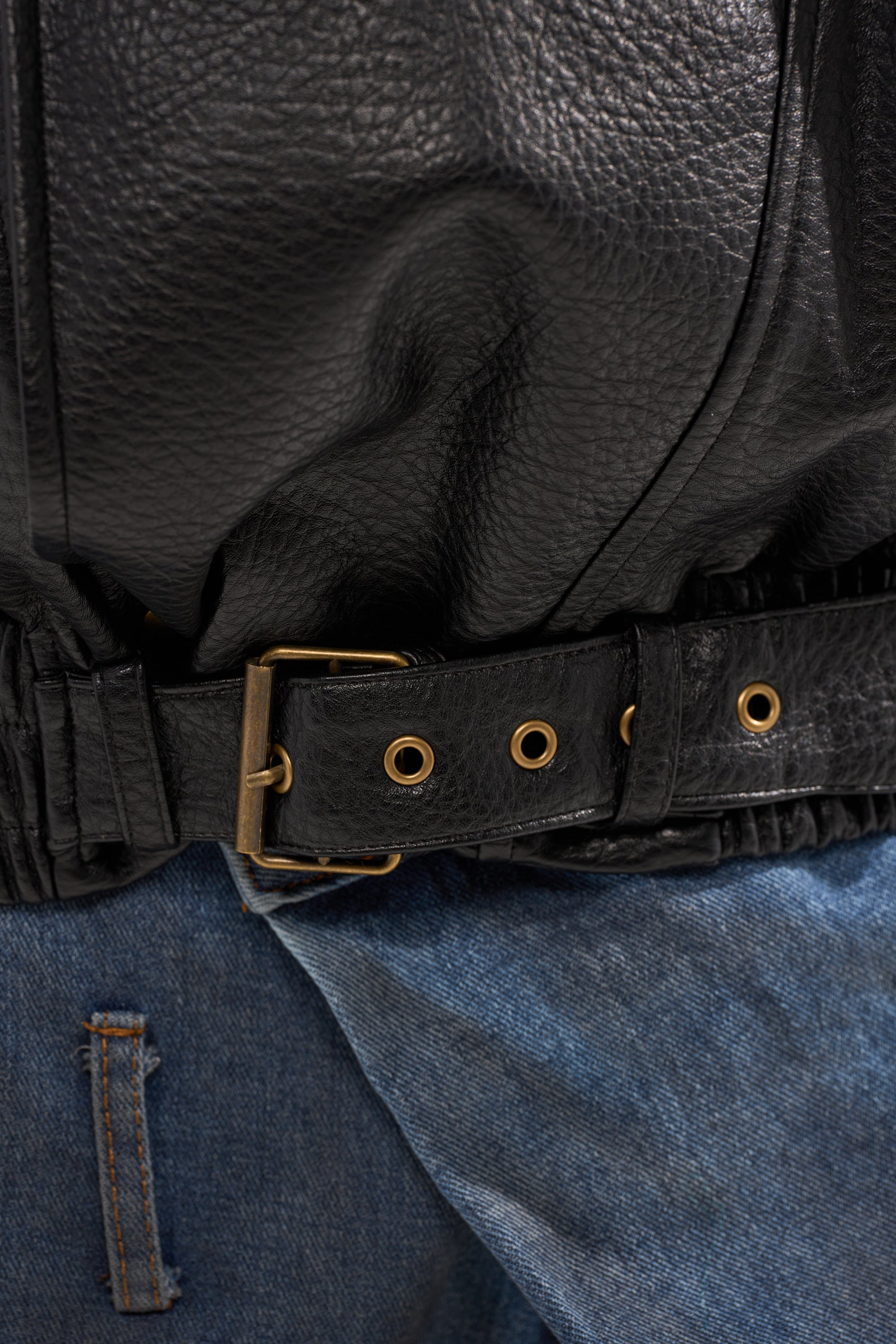 Close-up of the ERIKKA OVERSIZED MOTO black faux leather jacket with textured detail and a black belt with gold eyelets and buckle, styled with blue denim jeans for a streetwear-inspired look.