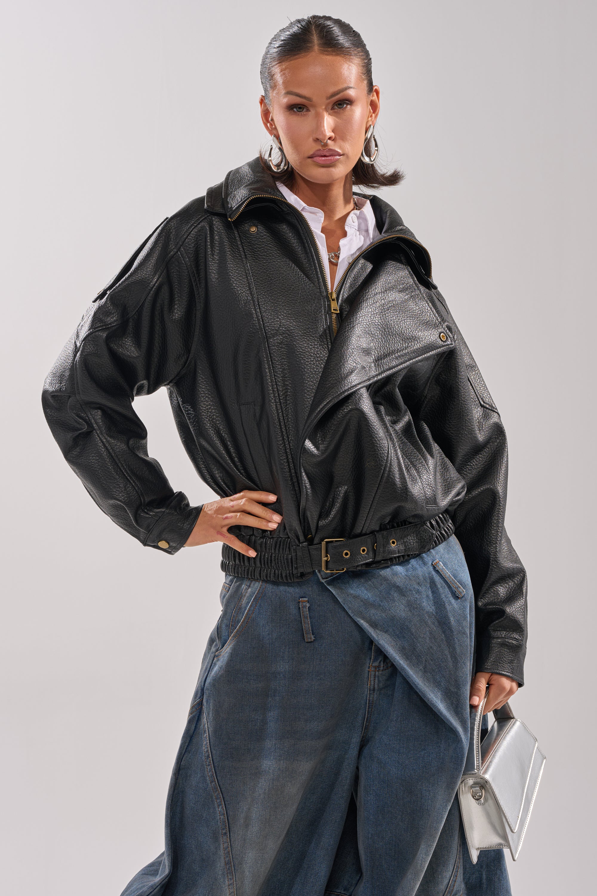A woman poses confidently in the ERIKKA OVERSIZED MOTO jacket with gold buttons and a belted waist, paired with a blue asymmetrical denim skirt, silver clutch, slicked-back hair, and hoop earrings.