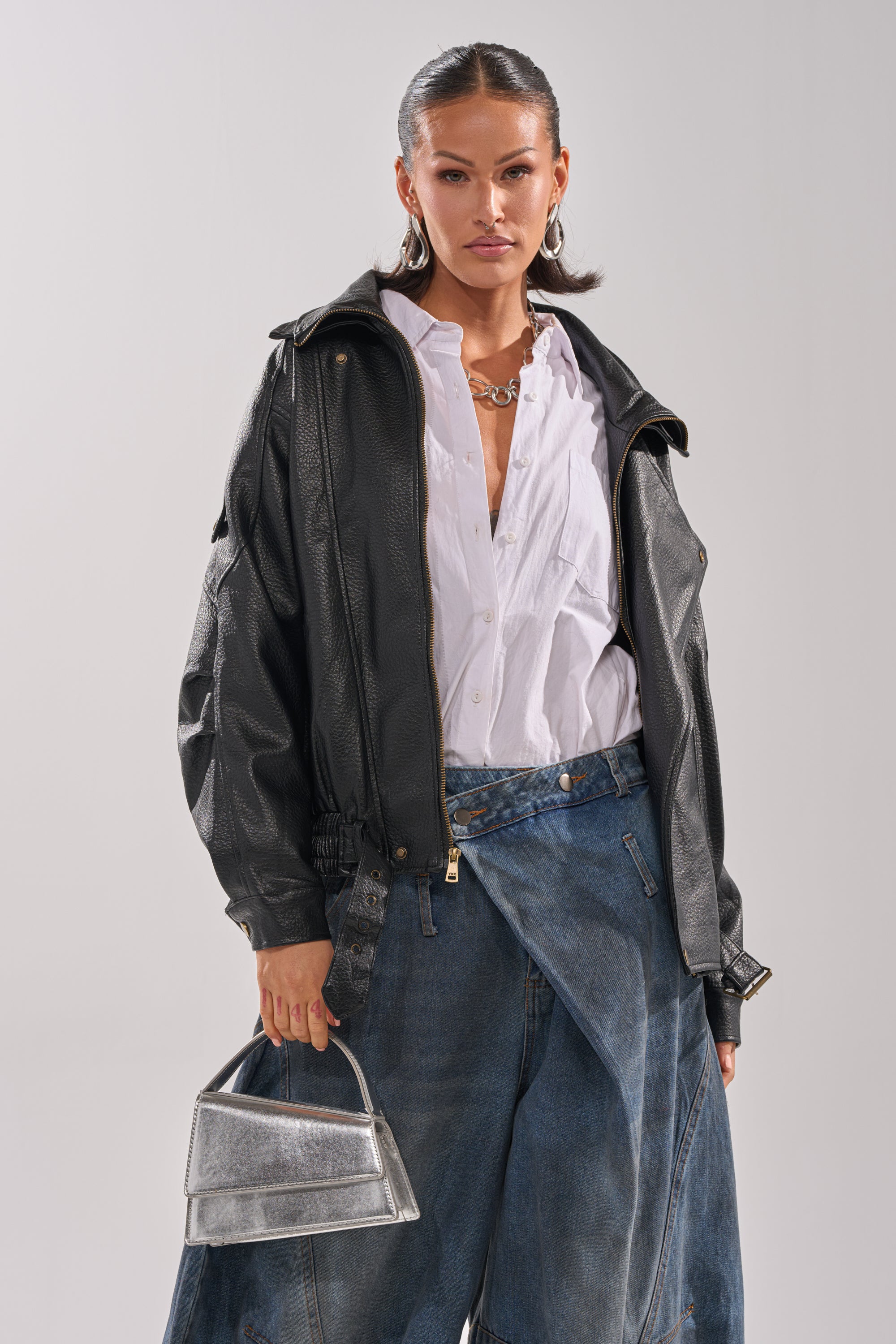 A woman confidently wears the ERIKKA OVERSIZED MOTO over a white shirt and wide-leg blue jeans, accessorized with gold jewelry, a small metallic silver handbag, and slicked-back hair.