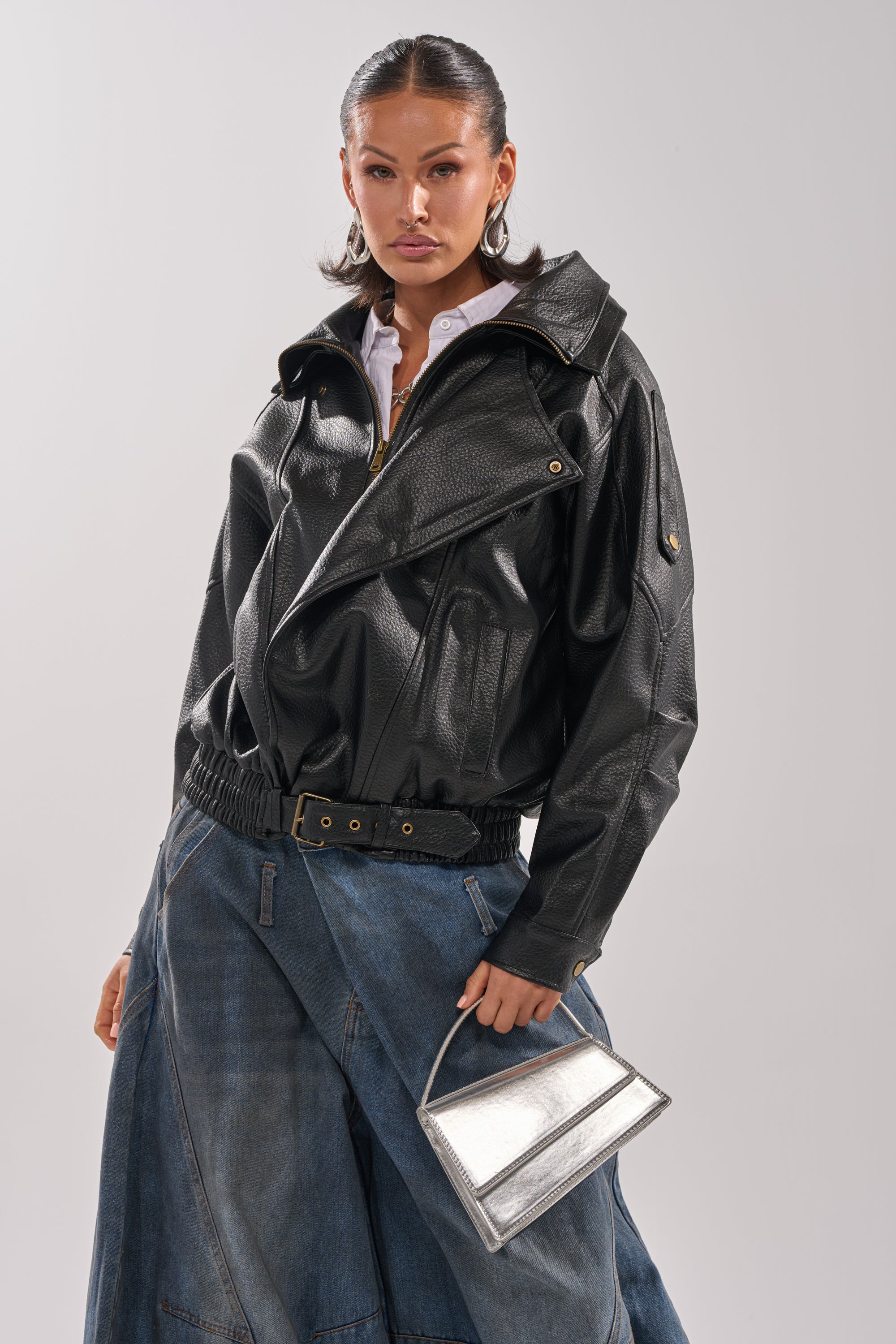 A woman with slicked-back hair and a confident look poses against a plain background in the ERIKKA OVERSIZED MOTO jacket, a white shirt, wide-leg blue jeans, and holding a small silver handbag.