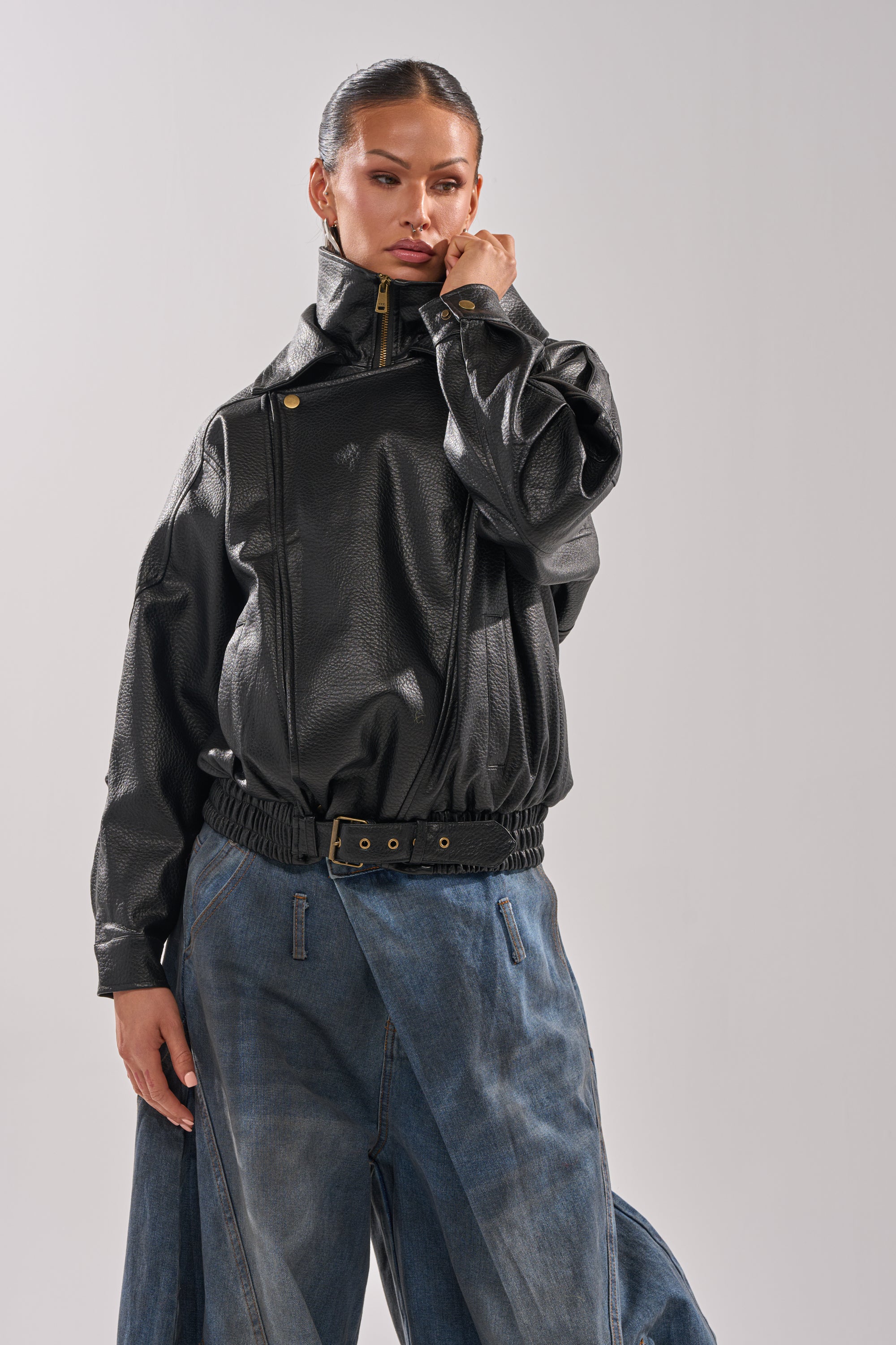A woman poses confidently in the ERIKKA OVERSIZED MOTO, a black moto jacket with a biker-inspired collar and belt, paired with loose blue denim pants against a plain light background.