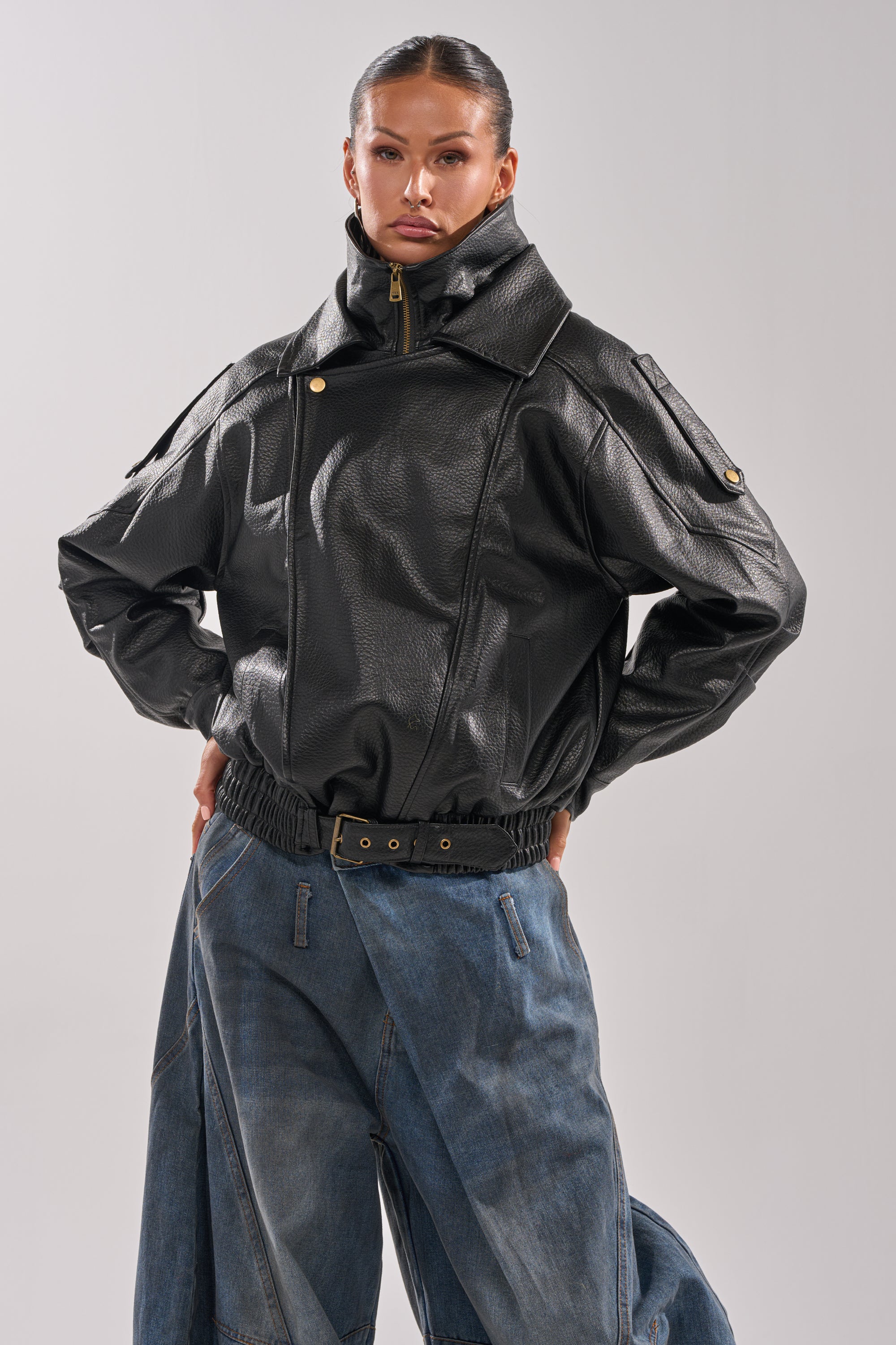 A person poses confidently in the ERIKKA OVERSIZED MOTO jacket, featuring a high collar and gold snap details, styled with loose blue jeans and a black belt against a plain light background.