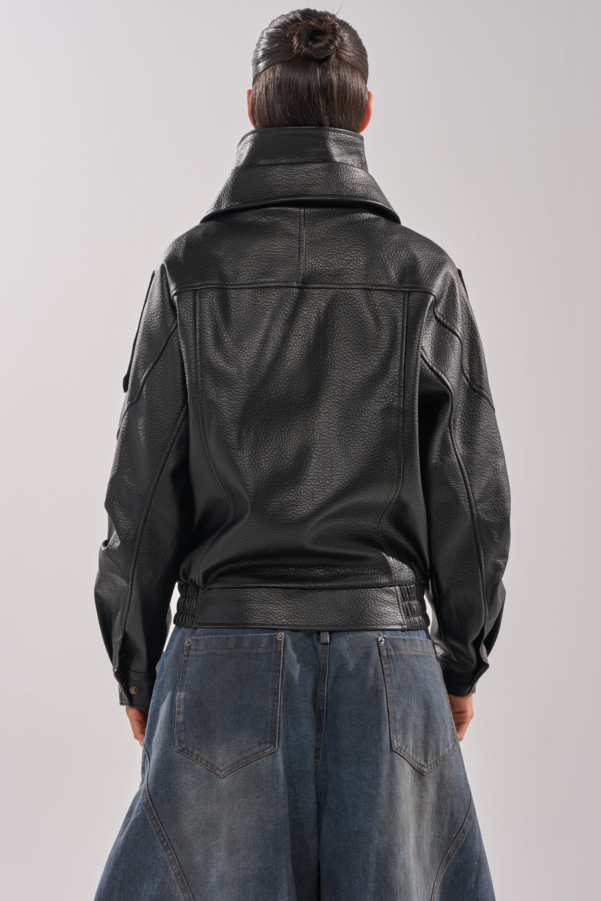 A person with dark hair in a bun is shown from behind wearing the ERIKKA OVERSIZED MOTO jacket and loose-fitting blue jeans with large back pockets.