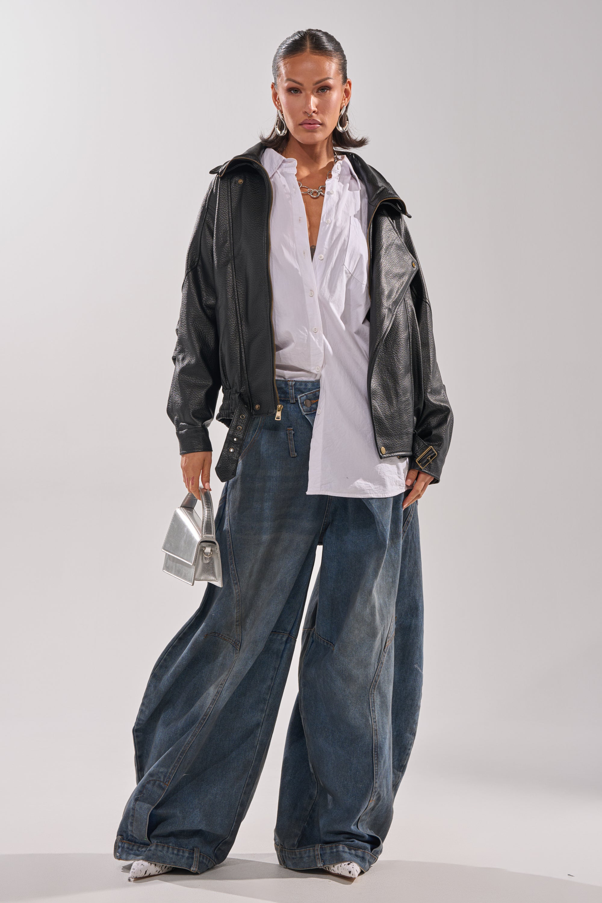 A woman poses against a plain background wearing the ERIKKA OVERSIZED MOTO jacket over a white shirt, wide-leg blue jeans, white sneakers, and holding a small silver handbag.