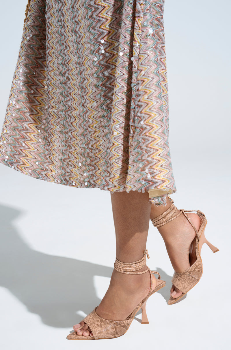 A person wearing a zigzag-patterned earthy-toned midi skirt stands on a light floor. Their legs and feet are shown with AKIRA OWENS TAN CHUNKY SANDALS, featuring brown textured open-toe high heels and ankle straps, while shadows create playful contrasts in the scene.