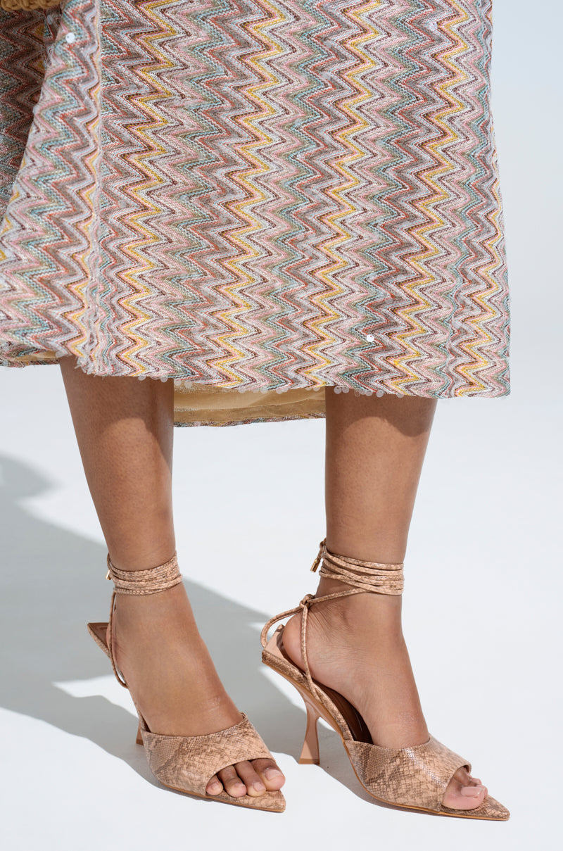 Close-up of a person wearing the AKIRA OWENS TAN CHUNKY SANDAL with ankle wraps, paired with a knee-length dress in a colorful zigzag pattern. The faux leather sandals perfectly complement the vibrant attire against a neutral background, highlighting the stylish legs and footwear.