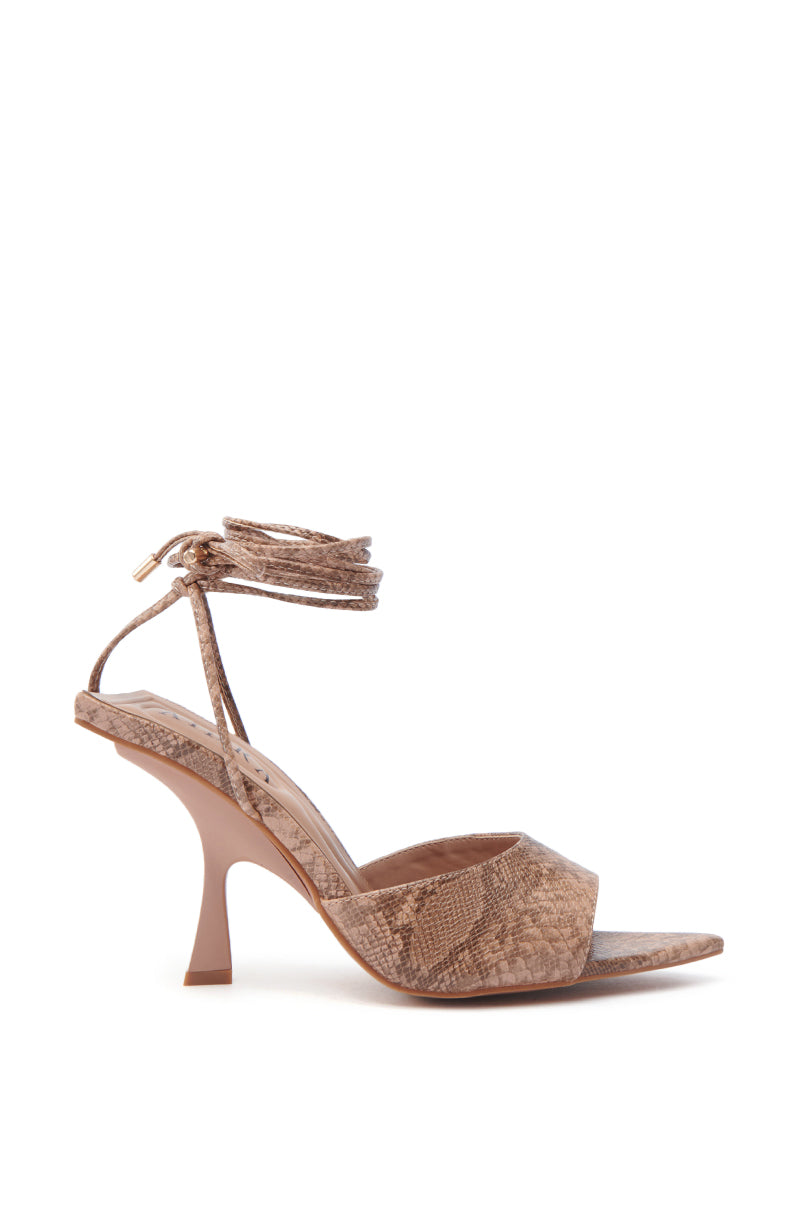 The AKIRA OWENS TAN CHUNKY SANDAL features faux leather with a textured surface and a closed toe strap. It includes a long, gold-tipped ankle strap elegantly wrapped around the ankle and boasts a thin stiletto heel, offering a refined design.