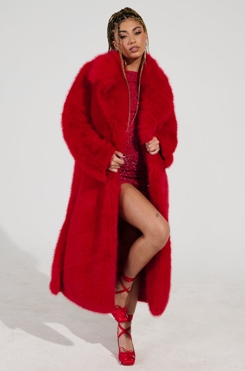 Dressed in a striking red OXFORD FAUX FUR TRENCH, its oversized design and satin-lined interior are perfectly complemented by their gleaming red dress underneath. With braided hair and matching red strapped high heels, they exude confidence against a simple white backdrop.