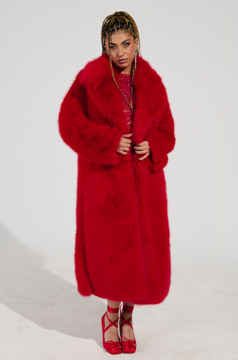 Confidently poised, a person dons an oversized maxi silhouette featuring the striking OXFORD FAUX FUR TRENCH in vibrant red. The coat's satin-lined interior complements a sparkly red ensemble paired with coordinated heels, while their hair is styled into braids against a plain backdrop.