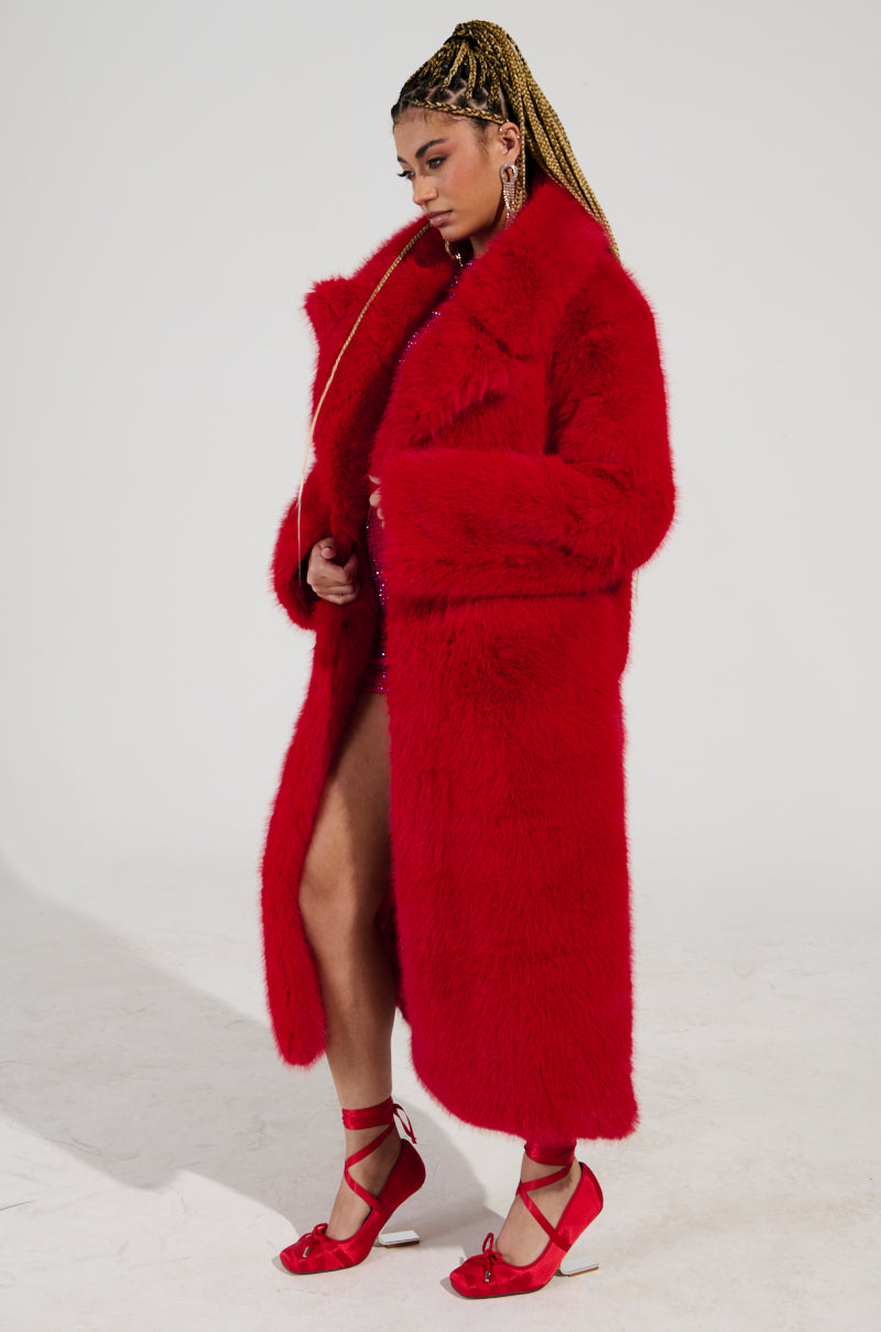 A person clad in a striking Oxford Faux Fur Trench, showcasing a long and fluffy red coat silhouette, poses against a simple backdrop. Their braided hair accentuates the outfit, with matching red shoes adding to the look. The open coat reveals hints of an ensemble underneath, featuring a satin-lined interior.