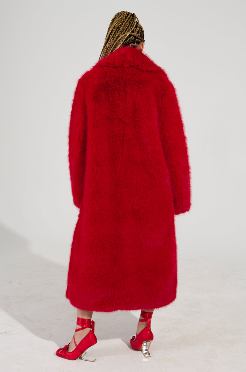 A person with braided hair stands with their back to the camera, elegantly wrapped in an OXFORD FAUX FUR TRENCH. The oversized maxi-length design complements red stockings and high heels adorned with silver details, all set against a plain white studio backdrop.