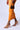A person in an orange skirt and OYSTA ORANGEMULTI SANDAL with abstract patterns strides confidently with a small leopard print handbag. They move gracefully across a plain white surface, highlighted by striking stiletto heels.