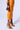 Dressed in a vibrant orange skirt, they're confidently holding a small leopard print bag and wearing OYSTA ORANGEMULTI SANDAL with stiletto heels. They exude style in a plain white studio setting.
