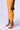 Against a white backdrop, a person models an orange fitted skirt and holds a small brown patterned handbag, showcasing the OYSTA ORANGEMULTI SANDAL with stiletto heels and purple accents for a playful touch.