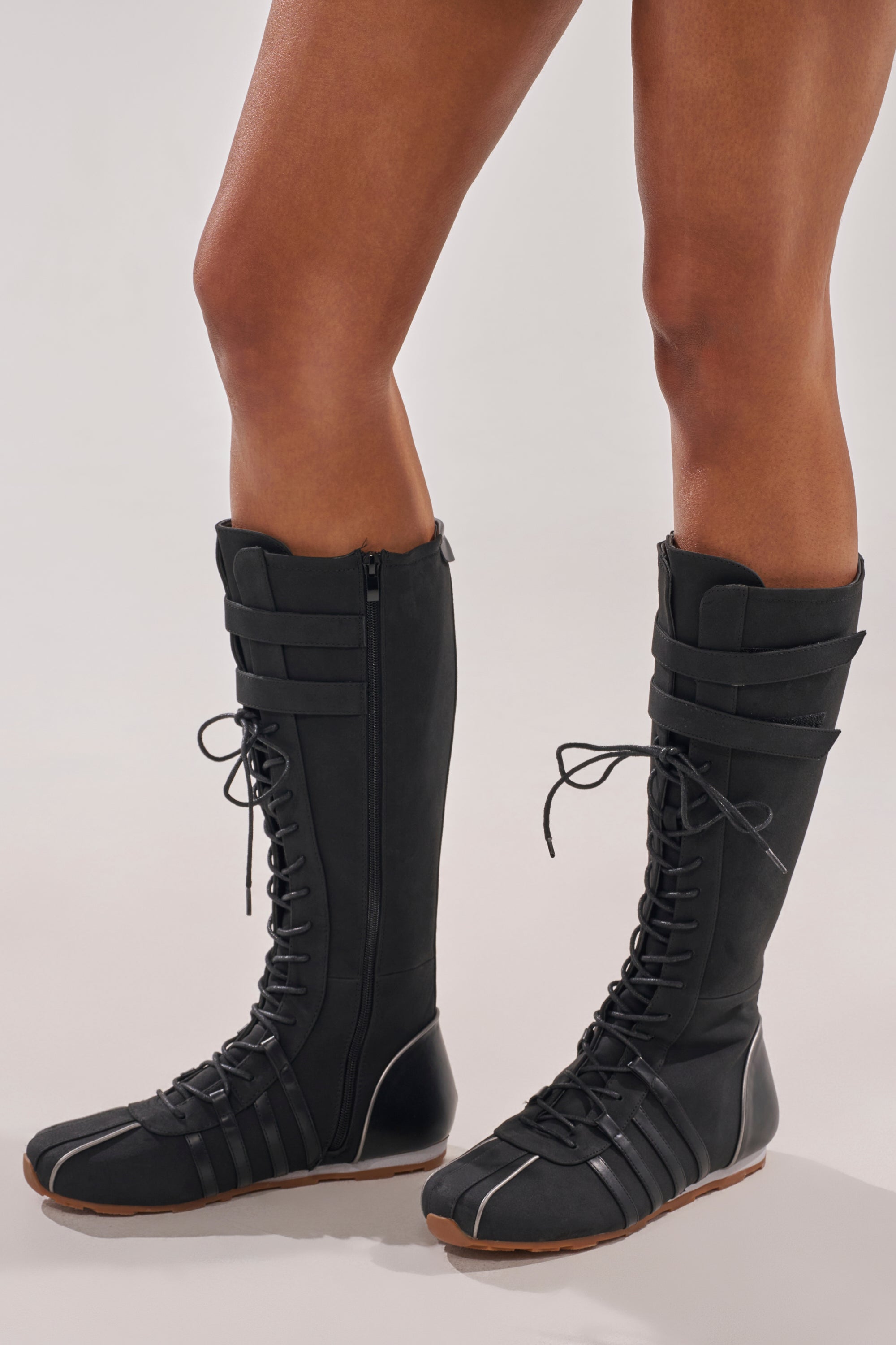 Wearing the AZALEA WANG OZZY BLACK FLAT SNEAKER—black, knee-high sneaker boots with lace-up fronts, Velcro straps, and brown soles—someone stands on a light surface. Only the sporty-chic legs and feet are shown. 0