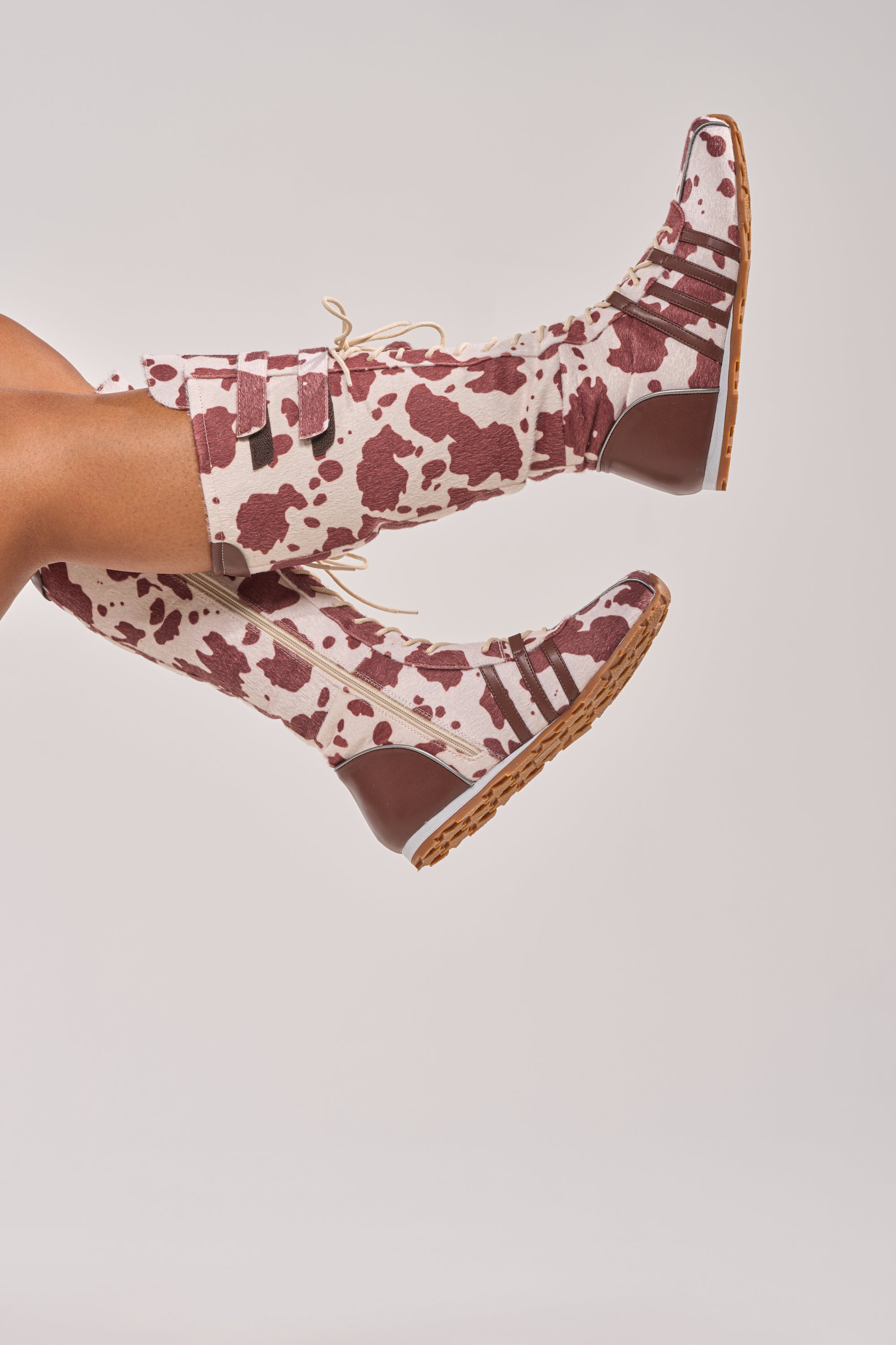 A person wearing AZALEA WANG OZZY BROWN MULTI FLAT KNEE HIGH SNEAKERS, featuring a brown and white cow print with brown soles, legs raised against a plain light background. 0